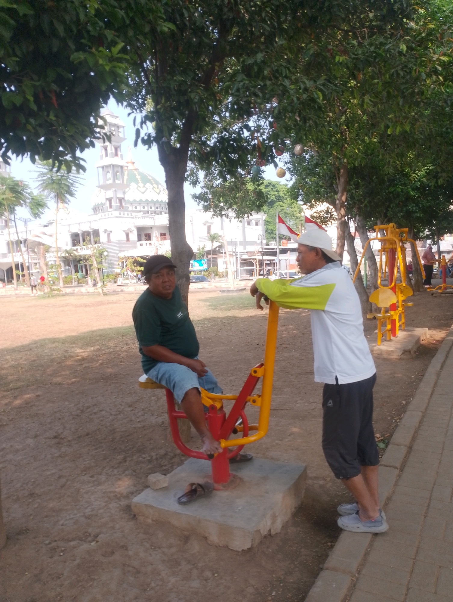 In Probolinggo, Indonesia, two adult males are present in an outdoor park setting, near a white building with a dome and minaret, likely a mosque. The ground is a mix of dirt and scattered leaves. To the left, a man wearing a dark green t-shirt and blue jeans sits on a red and yellow outdoor exercise machine. He has a dark baseball cap on and appears to be resting. To the right, another man in a white long-sleeved shirt with yellow accents on the sleeves, black capri pants, and grey slip-on shoes stands leaning on a similar red and yellow exercise machine. He is wearing a white cap backwards. In the background, several other pieces of yellow exercise equipment are visible, along with trees and a paved walkway. The lighting suggests it is daytime, with shadows cast on the ground.