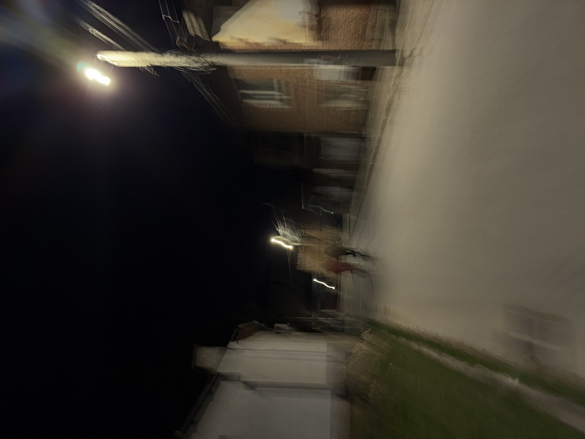 The image depicts an urban scene at night, potentially in Panagyurishte, Bulgaria. A street with a sidewalk is visible, illuminated by a street light. There is a building with brick facade and windows lining the street, and a green area of grass. A person wearing red can be seen on the sidewalk. The image has motion blur, which might be caused by a moving camera or moving objects in the scene.