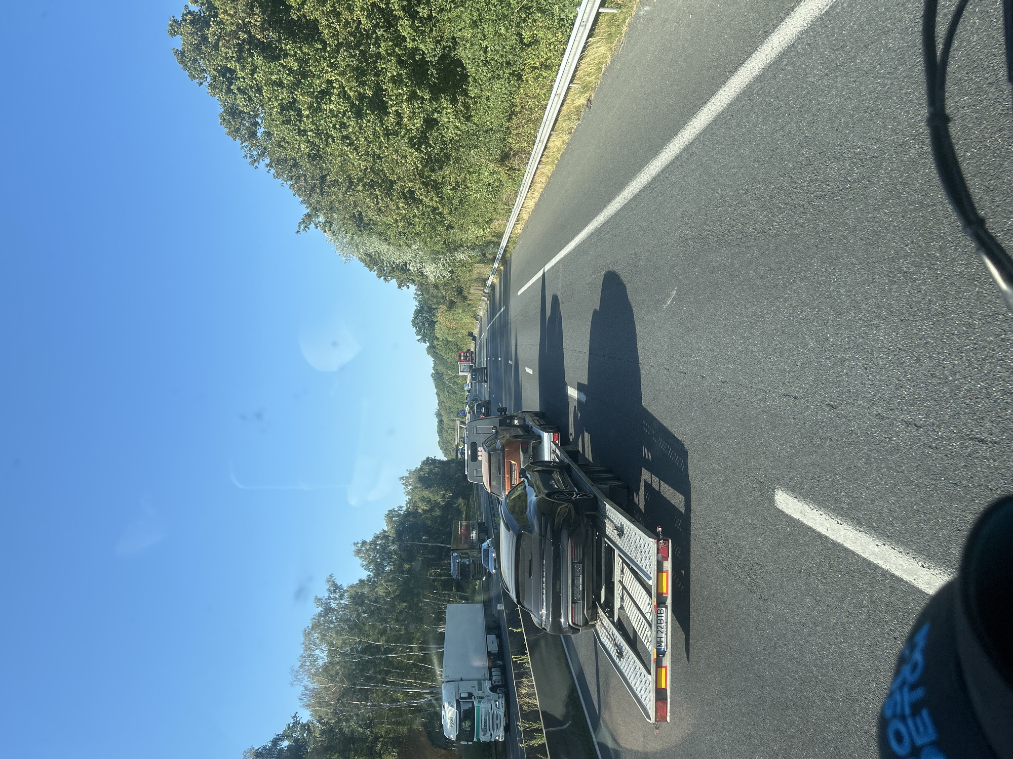 The image shows a highway in France with traffic. In the foreground, a car carrier trailer is visible, carrying a black Porsche. Behind it, an orange SUV is also on the trailer. The highway is bordered by trees on the right and left. The sky is clear and blue. The location is identified as Thiers-sur-Thève, France.