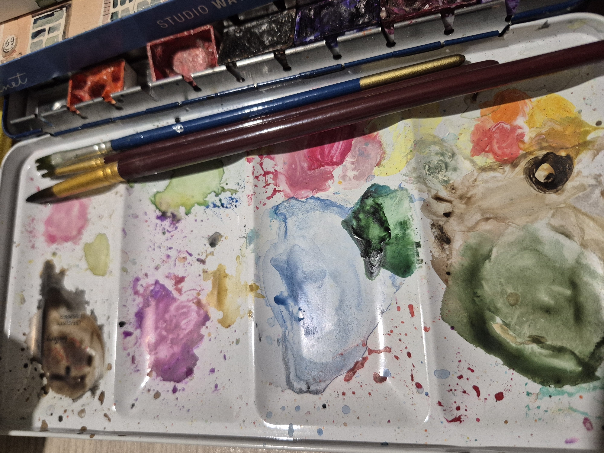 The image displays a well-used watercolor palette with various colors mixed and splattered across its surface. Several paintbrushes, with wooden handles in shades of burgundy, gold, and blue, rest on the palette. To the right of the palette is a metal tin containing rows of watercolor pans, some of which appear to be filled with paint. The overall impression is one of a creative workspace, likely used for artistic endeavors. The image does not contain any discernible elements that would place it in the city of Popovo, Bulgaria, or any specific urban environment.