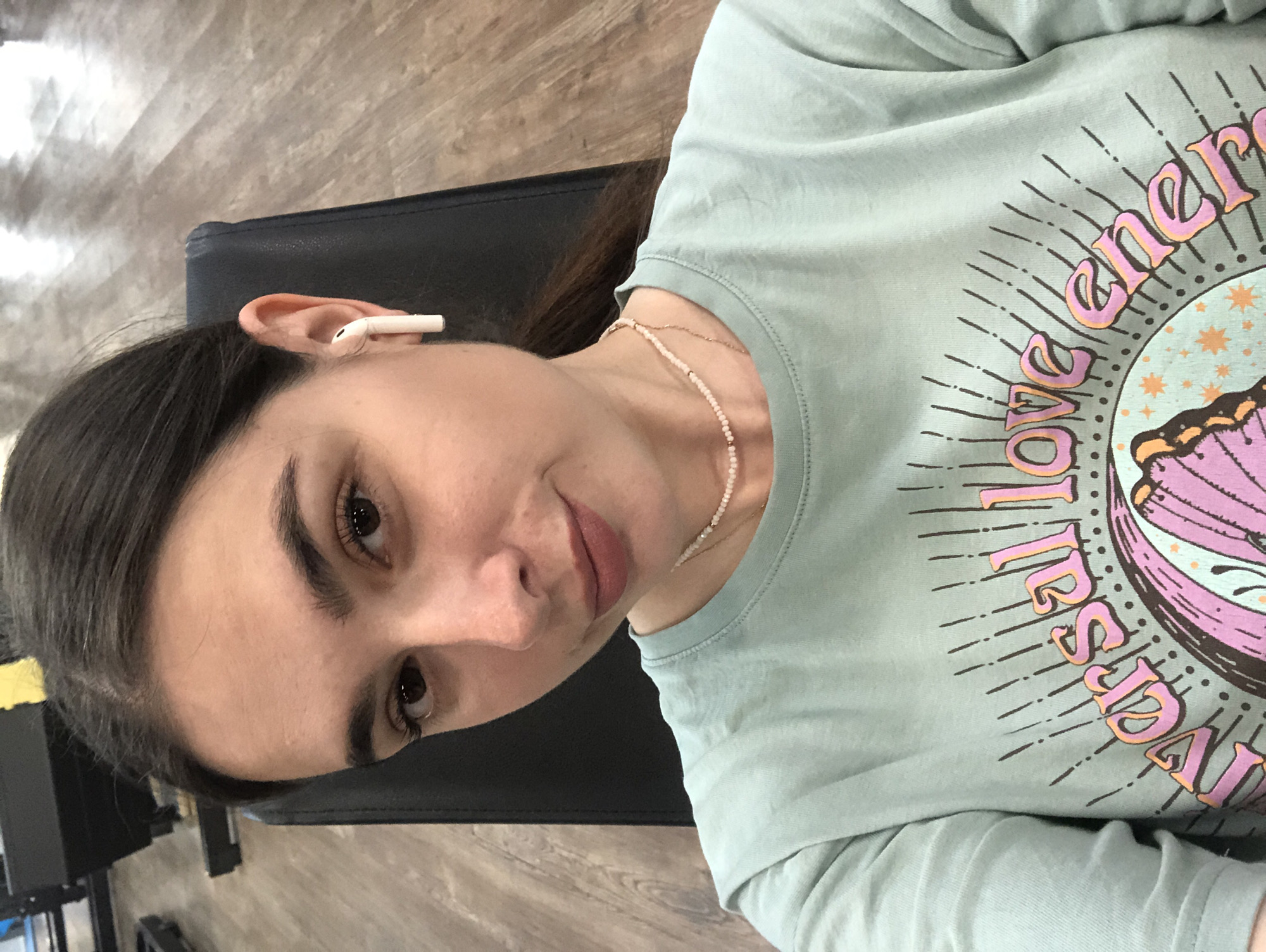 In a selfie taken from a slightly high angle, a young woman with dark hair pulled back is shown from the chest up. She is wearing a light blue t-shirt with a graphic design that includes text and an illustration of a sun with a butterfly. She has on a delicate beaded necklace. A white wireless earbud is visible in her right ear. The background suggests an indoor setting, possibly a gym, with dark seating and a wooden floor. The woman is looking towards the camera with a slight smile. The scene is indoors.