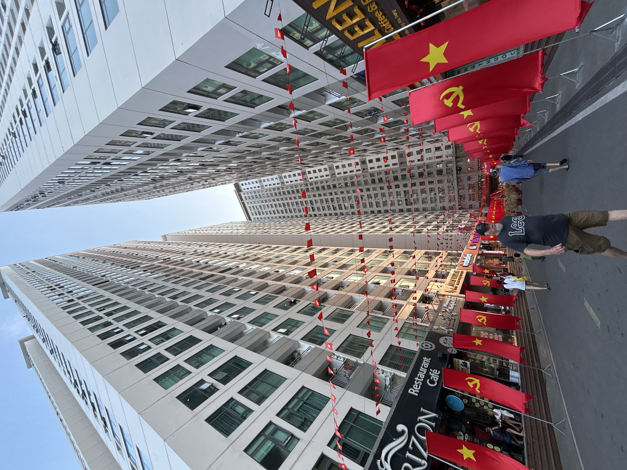 The image depicts a bustling urban street scene in Bắc Nha Trang, Vietnam, flanked by two towering, modern high-rise buildings. The perspective is from street level, looking up between the buildings, emphasizing their height and the overhead decorations.
The street is extensively decorated with numerous red flags. Large rectangular flags, featuring either a yellow star (the National Flag of Vietnam) or a yellow hammer and sickle (symbol of the Communist Party), are mounted on poles lining both sides of the thoroughfare. Above the street, long strings of smaller red flags, each with a yellow star, are strung across, forming a festive canopy between the buildings.
Two men are visible on the street. In the foreground, a man wearing a dark t-shirt with "LEE" emblazoned on it, khaki shorts, and a cap stands facing towards the right side of the frame. Further down, another man in a blue shirt and dark shorts walks away from the camera. The ground floors of the buildings feature various commercial establishments, including a "Restaurant Café" with "RIZON" visible on a prominent black sign. Other signs, some with Vietnamese text, suggest additional businesses like coffee shops.
The high-rise buildings are uniformly light-colored, possibly white or light gray, with numerous rectangular windows, some of which are open. The architecture appears modern and functional. The clear sky and bright, even illumination suggest it is daytime, likely late morning or early afternoon. The pervasive display of flags indicates a celebratory atmosphere, possibly for a national holiday or significant event in Vietnam.