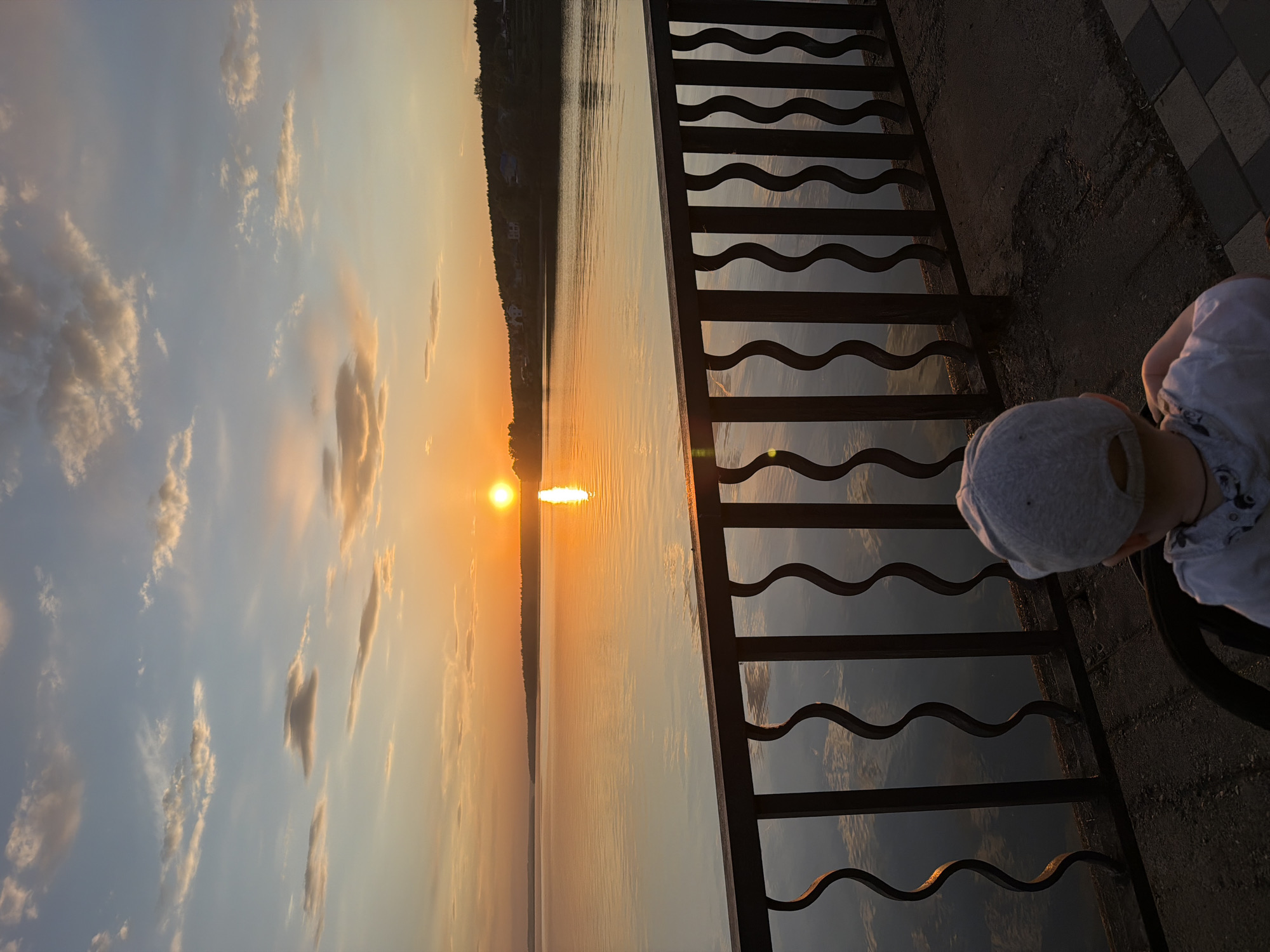 Sunset over the Votkinsk Reservoir in Votkinsk, Russia. A person wearing a grey cap and white clothing is near a dark metal railing. The water reflects the orange and yellow light from the setting sun, which is visible on the horizon. Light clouds are scattered in the blue sky. Land is visible on the opposite shore of the reservoir.