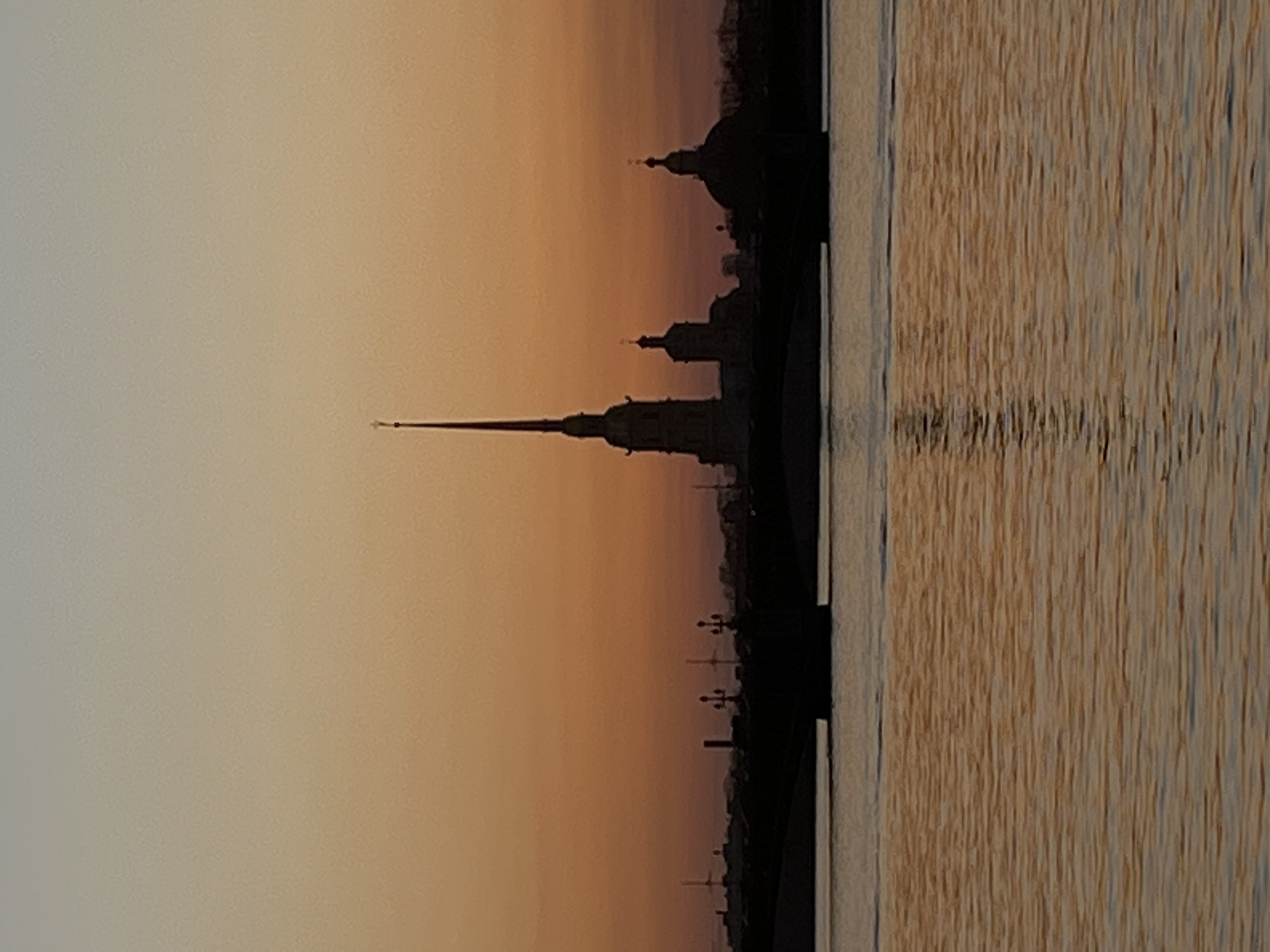 The image depicts a skyline of Saint Petersburg, Russia at sunset, reflected in the Neva River. Silhouetted against the warm tones of the sky are the Peter and Paul Cathedral and other prominent buildings. The river's surface captures the light and form of the cityscape in a blurry, horizontal reflection. The photograph is taken from a low angle, close to the water level, enhancing the reflection.