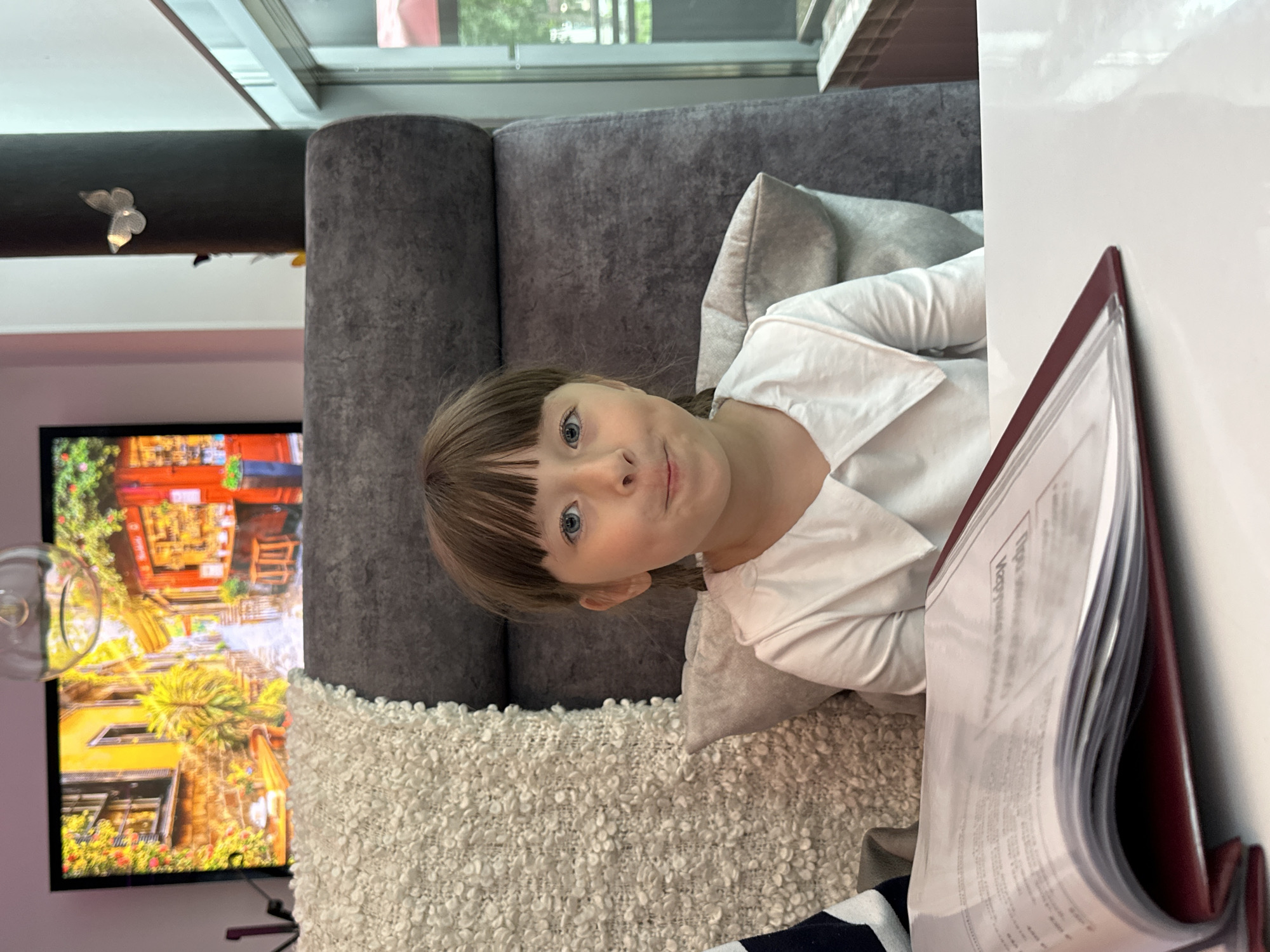A young girl with blonde hair and blue eyes is lying on a couch, looking at the camera. She is wearing a white collared shirt. A large grey pillow is behind her head. The couch appears to be in a living room, with a television screen visible to the left displaying a colorful image of a street scene with buildings and greenery. A textured white pillow is on the couch in front of the television. To the right of the girl is a burgundy binder or folder filled with papers, partially obscuring the edge of a white surface, possibly a table. A window with a view of trees and buildings is in the background, suggesting it is daytime. The overall scene appears to be relaxed and casual.