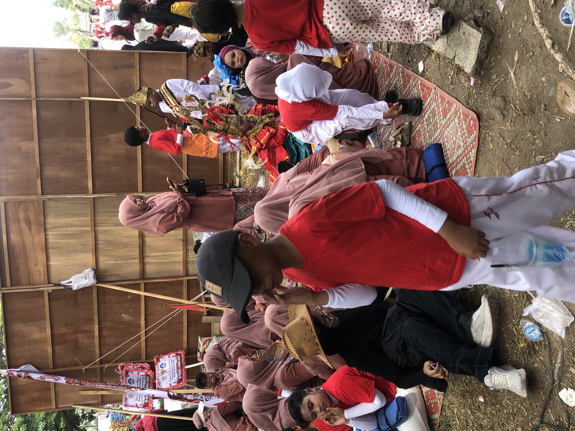 This is a medium shot, eye-level, full-color outdoor photograph taken in Lhokseumawe, Indonesia. The scene depicts a gathering of people, primarily children and adults, in what appears to be a festival or cultural event. The backdrop is a large, wooden wall constructed from planks, supported by wooden poles. To the left, a tall banner with red and white stripes and text is attached to one of the poles, alongside smaller square signs with red borders.
In the foreground, a young boy wearing a red t-shirt and white pants with red accents is the most prominent figure. He has a black baseball cap on and is looking down, with his right hand touching his forehead. To his left, another boy in a red t-shirt with white sleeves is seated, looking towards the left of the frame, his hands clasped in front of him. A man in a black t-shirt and black pants is seated behind him, wearing a tan cowboy hat.
The middle ground is populated by a group of individuals, many of whom are wearing traditional Indonesian clothing, including headscarves in shades of pink and brown. One woman in a pink headscarf and brown dress is standing and looking at her phone. To her right, a young boy in a red shirt and orange sash stands next to another boy wearing a gold crown and a white and red ornate garment. Further back, a young person in a bright red and gold costume is seated. Other individuals are visible in the background, including some in red shirts and white polka dot pants.
The ground is covered with a woven mat that has a red and green diamond pattern. There are various objects scattered on the ground, including plastic bottles, plastic bags containing items, and a blue cylinder. The overall environment appears to be a dusty, outdoor area.