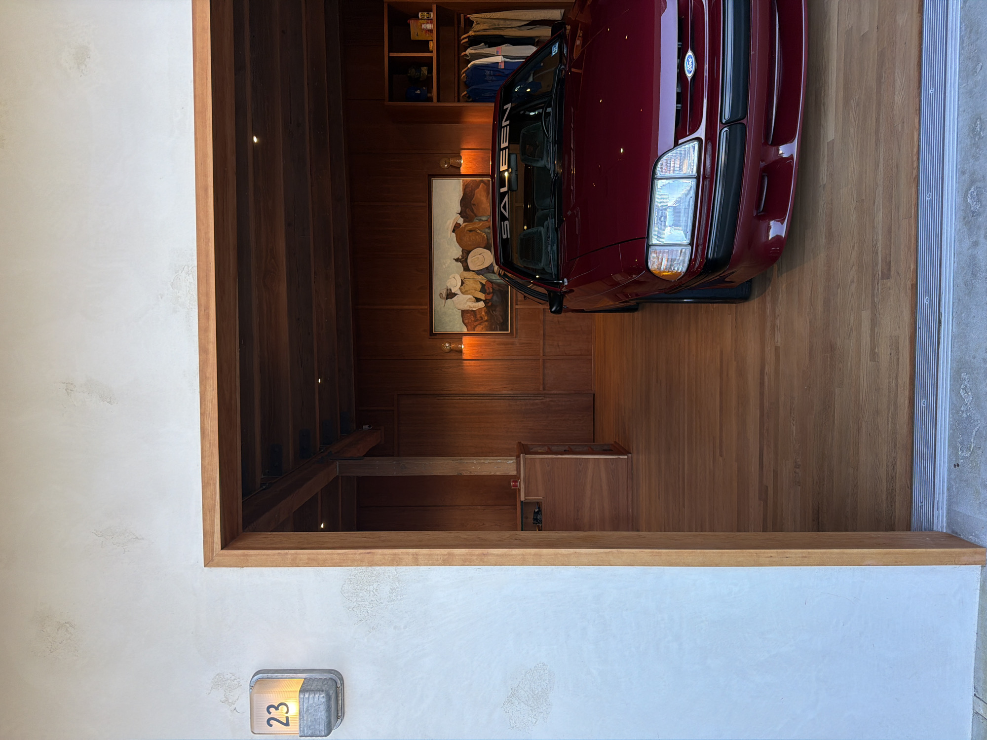 In Malibu, California, a red Ford Mustang Saleen is parked inside a garage with wooden walls and flooring. The car, which appears to be from the late 1980s or early 1990s, has a "SALEEN" decal across the top of its windshield. On the wall behind the car is a framed painting depicting three cowboys. To the right of the painting, a closet area is visible with clothes hanging on racks. The garage is accessed through a wide wooden doorway, and an illuminated outdoor light fixture displaying the number "23" is mounted on the exterior wall to the left of the entrance. The overall scene suggests a well-maintained garage or showroom.