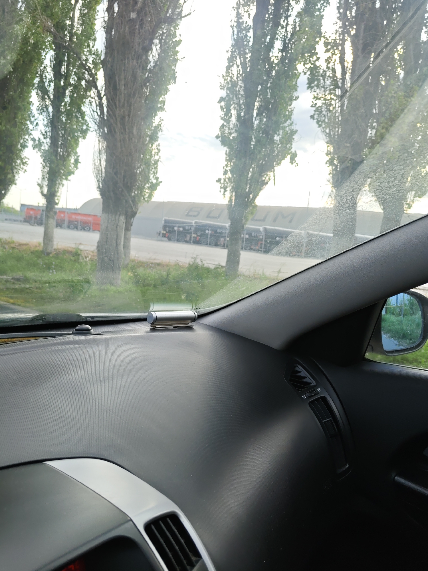 This image is a view from inside a car, looking out through the windshield. The dashboard is in the foreground, with a silver air freshener and a black sensor visible. Outside the car, there are several tall, slender trees with green foliage and bare branches. Beyond the trees, a large building with the letters "BUDM" visible on its facade is present. In front of the building, several red trucks and what appear to be black trailers are parked. The scene suggests an industrial or commercial area, possibly a logistics hub or factory. The sky is bright and overcast, indicating daytime. The image is undated and unsigned. It is not possible to definitively determine the exact location within Novocherkassk, Russia from the image alone, but the presence of industrial structures and the style of vehicles might be indicative of such a setting.
