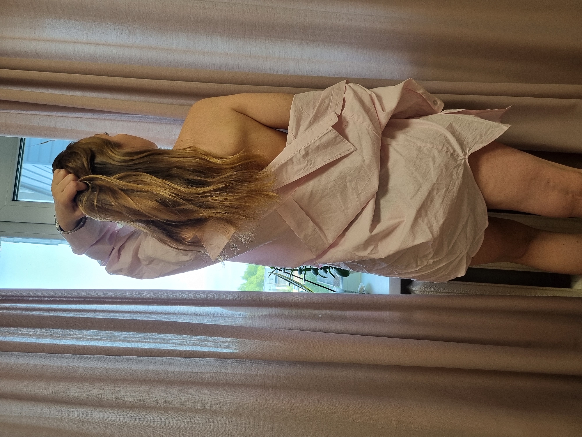 A fair-skinned person, possibly female, with long, light brown hair, is lying in a bed near a window. The person is wearing a light pink, oversized shirt. The left hand is near their head. Outside the window, plants and buildings are visible. The image is taken indoors, with curtains in the background. The location is not identifiable as Chaikovsky, Russia from the given image.