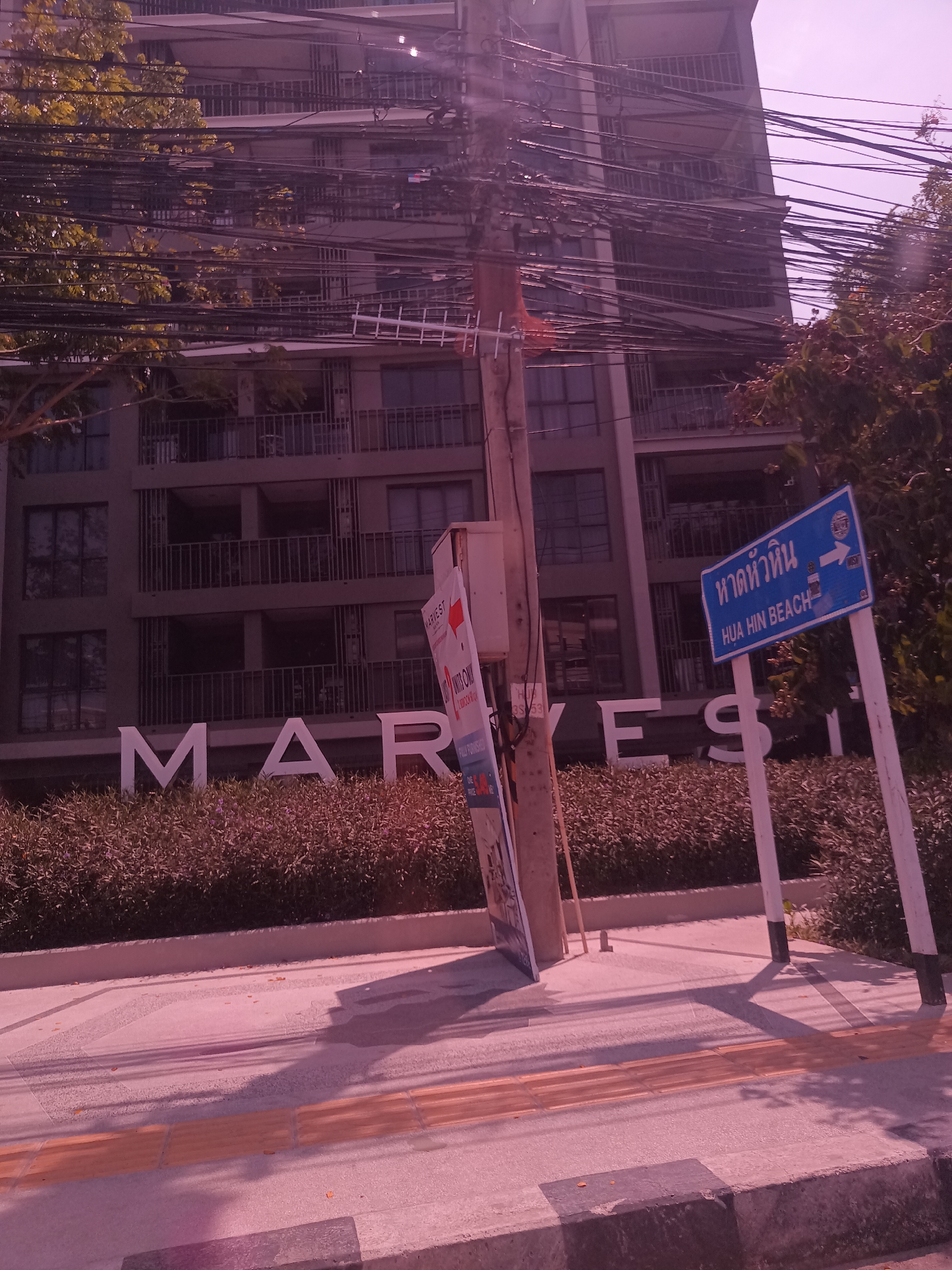 The scene is located in Hua Hin City Municipality, Thailand. It depicts a building with large white lettering "MARVES" in front of a low hedge. A blue sign reads "Hua Hin Beach" in Thai and English with an arrow pointing right. There are power lines above and a utility pole to the left of the sign. A promotional advertisement is attached to the utility pole. A paved road with tactile paving for the visually impaired is in the foreground.