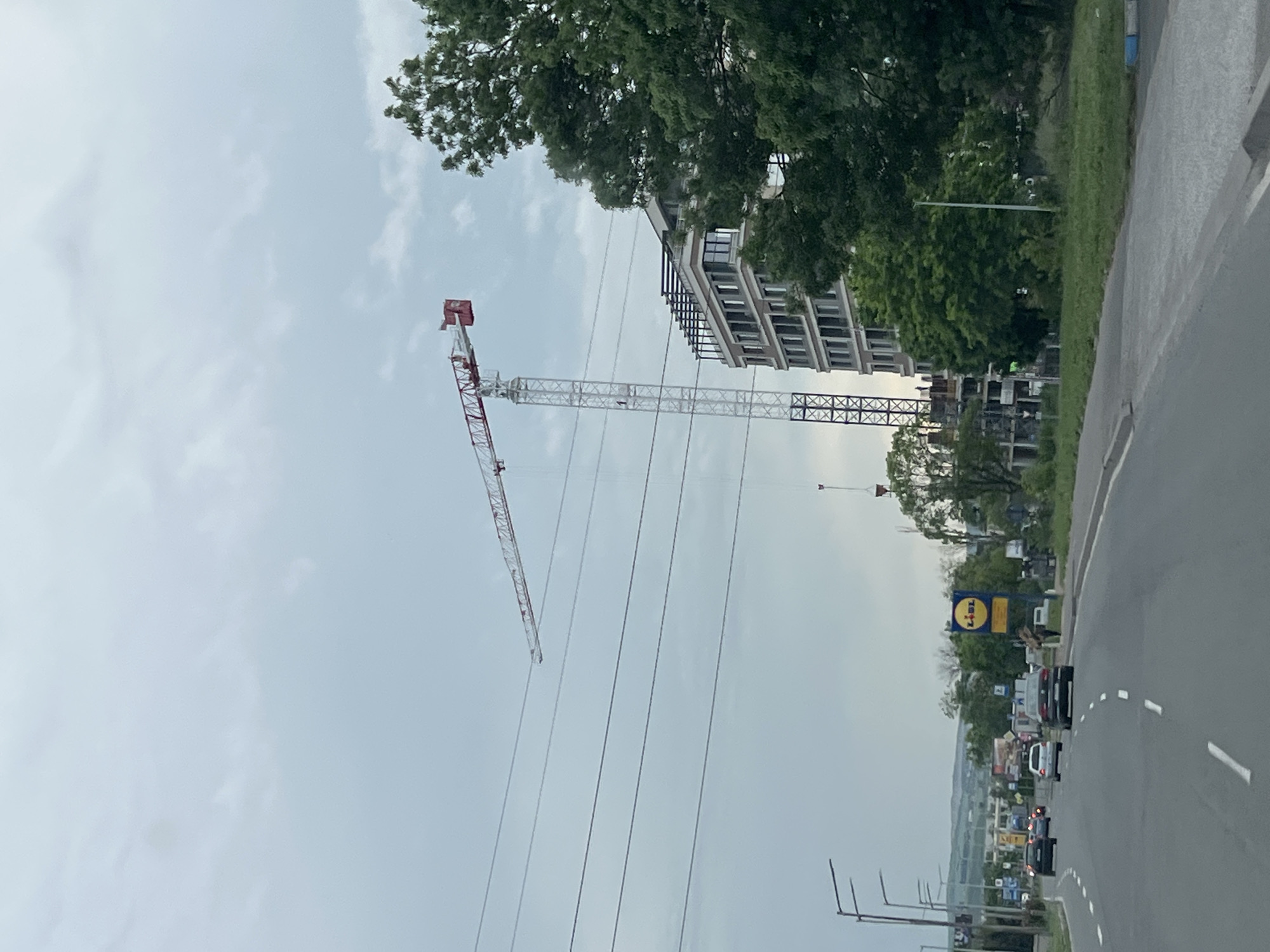 The image shows an outdoor scene in Burgas, Bulgaria, depicting a street with a construction site in the background. Prominent in the frame is a large white crane with a red jib, positioned near a multi-story building under construction. The building has a visible framework of concrete and steel. Overhead power lines are visible, stretching across the scene. To the right, a road is seen with intermittent white lane markings and a few cars driving along it. In the distance, a Lidl supermarket sign is visible amidst the street. The sky is overcast, casting diffused light over the scene. Trees line the right side of the road, adding greenery to the urban environment.