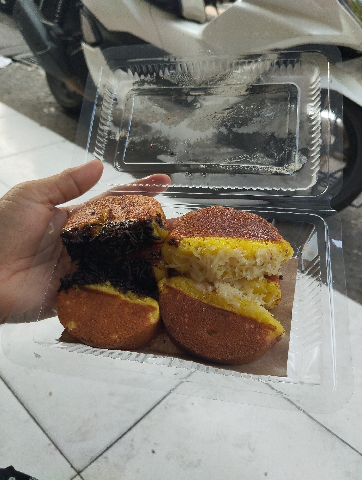 A hand is holding a clear plastic container filled with sweet martabak, a type of pancake. Two pieces of martabak with chocolate sprinkles are stacked on the left, and two pieces with grated cheese are stacked on the right. The container is open and is placed on a tiled surface. In the background, a white motorcycle is partially visible, suggesting an urban street setting, possibly in Surabaya, Indonesia, given the style of the martabak and the common practice of purchasing street food there. The lighting indicates it is daytime.