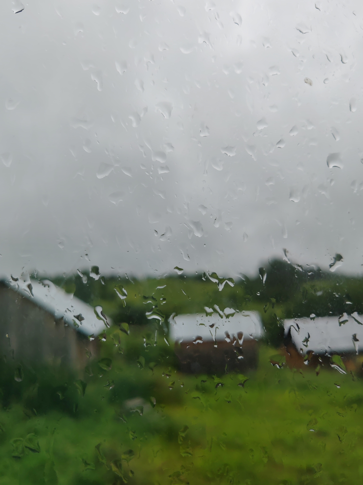 The image shows a view through a window covered in raindrops. Outside, there are several small, simple structures, possibly sheds or houses, with dark roofs and light-colored walls, set against a backdrop of rolling green hills under a cloudy, overcast sky. The rain obscures the details of the buildings and the landscape, creating a soft-focus, melancholic atmosphere. The scene appears to be in a rural or semi-rural setting.