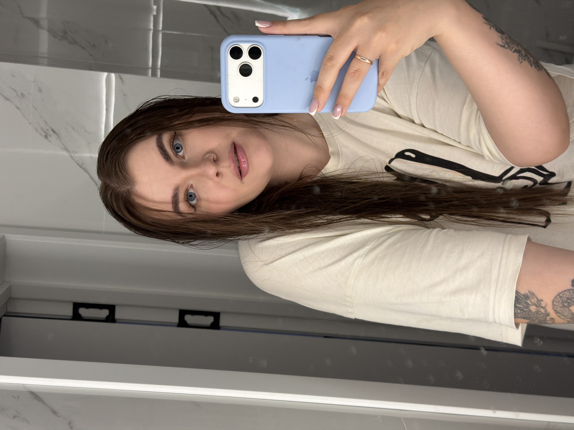 A young woman with long brown hair takes a selfie in a bathroom. She is wearing a light-colored t-shirt and has a ring on her right hand. Her phone, with a light blue case, is visible in her hand. The background shows a tiled wall and a door frame. There are no specific details in the image that indicate the location is Chaikovsky, Russia.