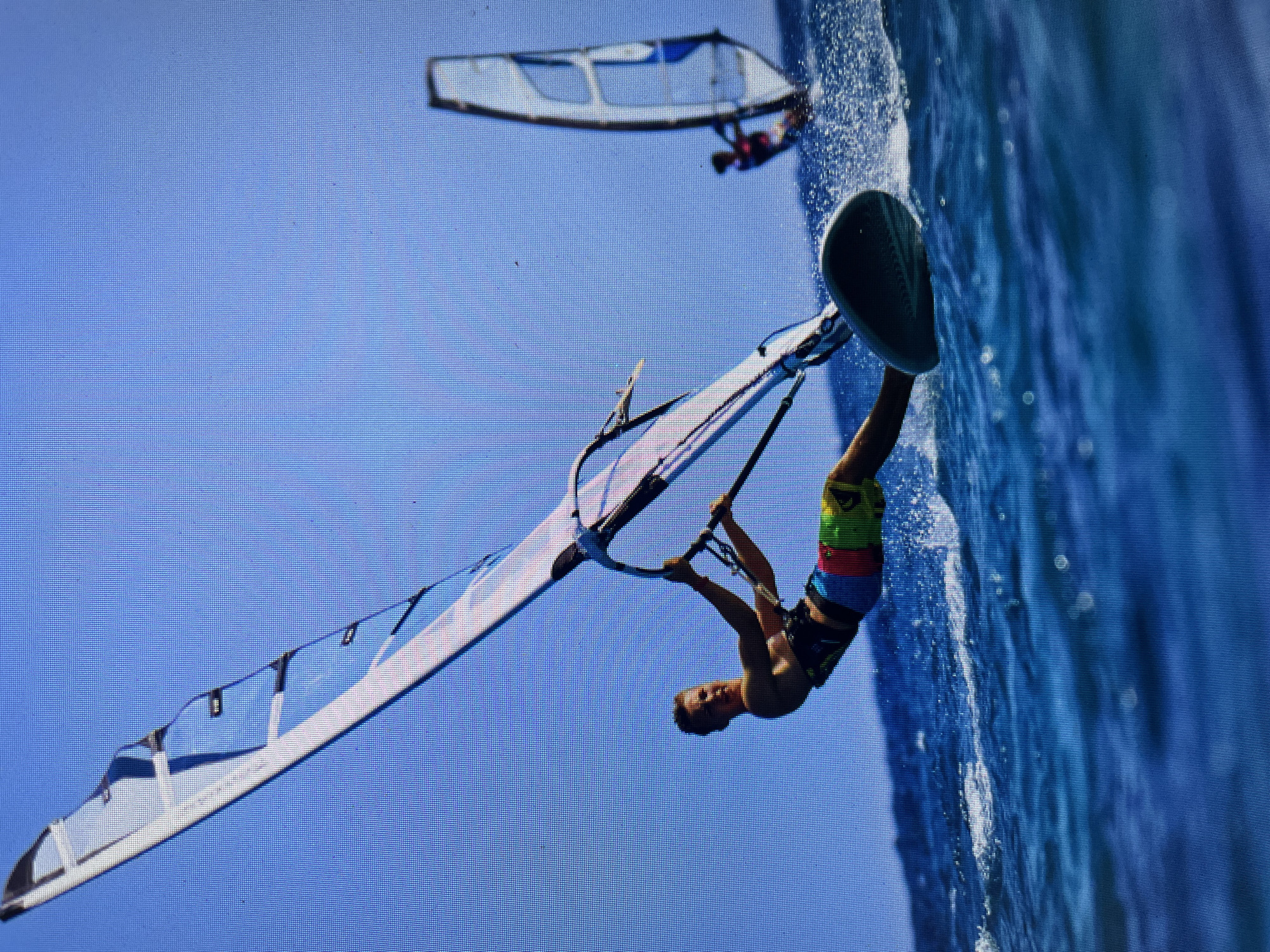 The image shows a windsurfer performing a trick in the water. The windsurfer is upside down, holding onto the boom of the sail with both hands. The sail is white with blue accents. The board is dark green. The water is blue and choppy. In the background, another windsurfer is visible in the distance. The location is possibly in the city of Bacong, Philippines.
