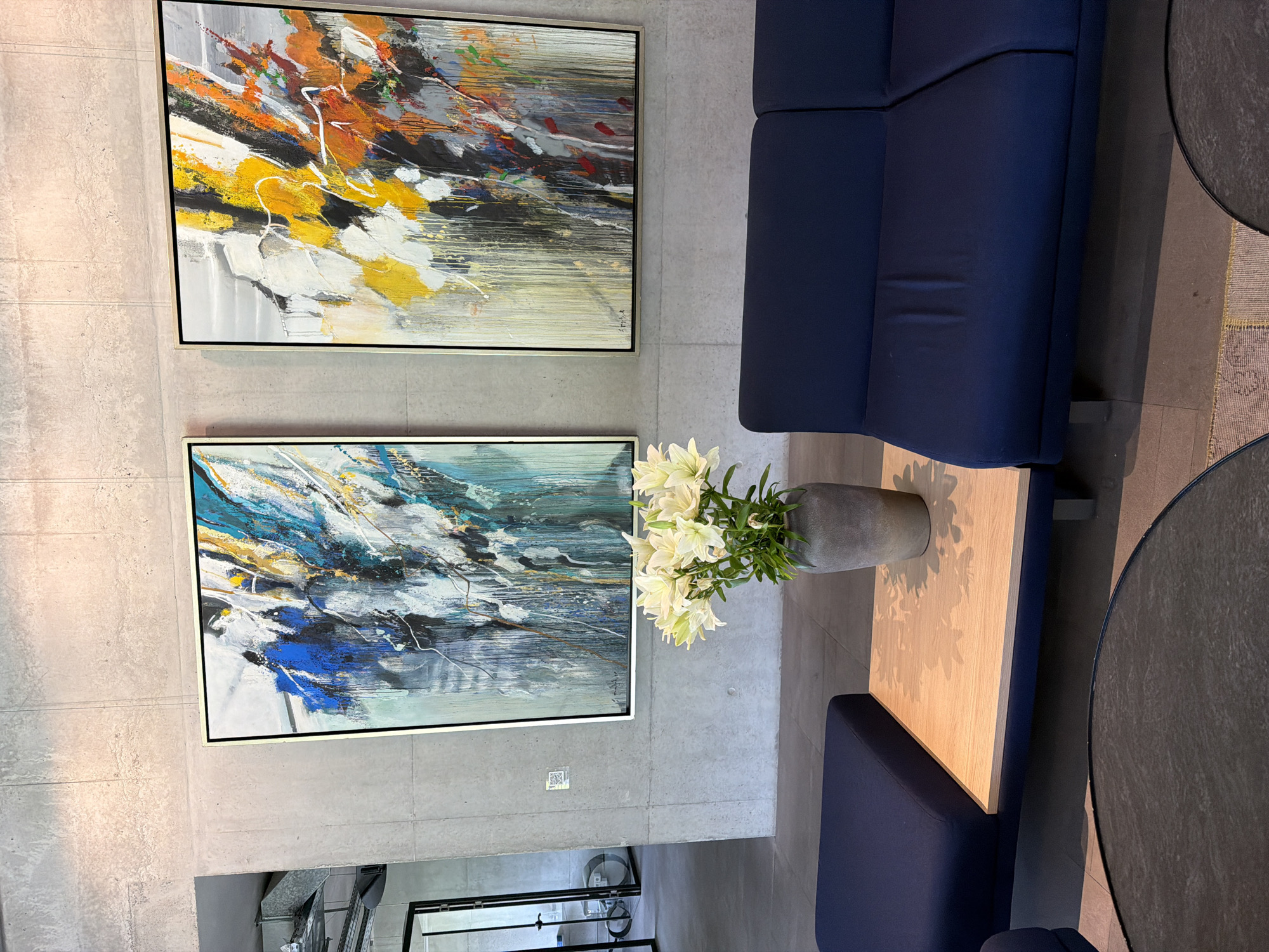 The image shows an indoor scene with two abstract paintings hanging on a gray wall, a navy blue sofa, a small wooden table with a gray vase containing white lilies, and two round black tables. Below the paintings are visible parts of a metal structure, perhaps part of a rack or stand. The location is Chkalov, Armenia, given the prompt.