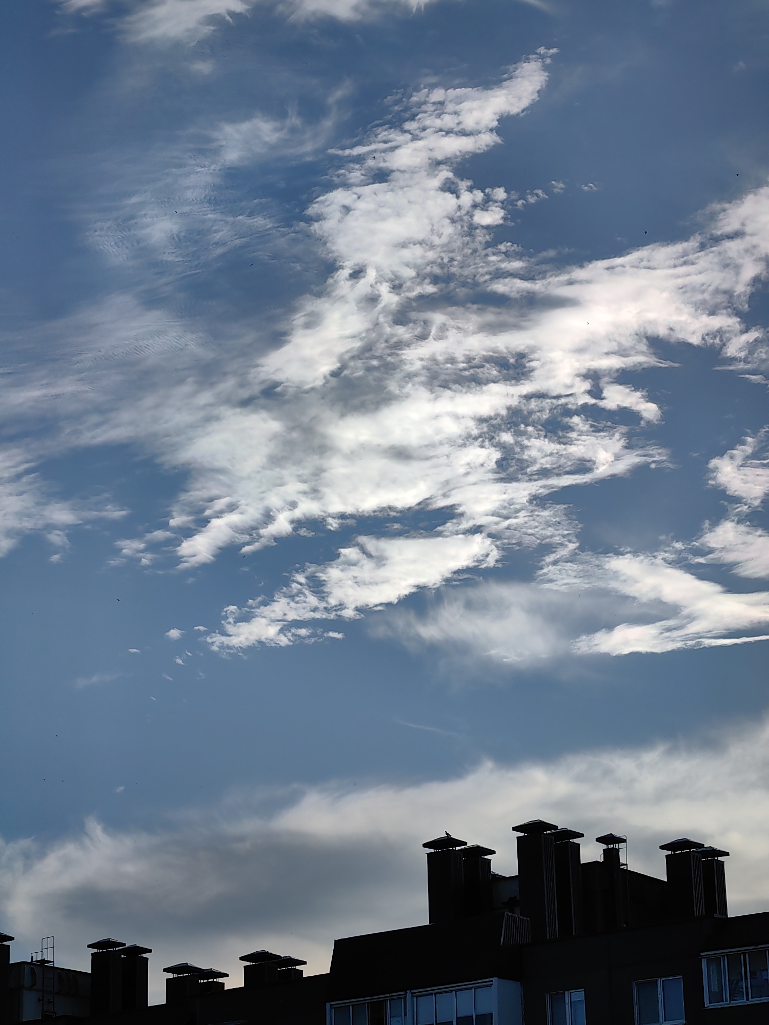 The image depicts a silhouetted view of apartment building rooftops with numerous chimneys against a partially cloudy sky. The buildings are dark, with some windows visible, suggesting an urban or suburban setting. The sky is a mix of blue and white, with wispy clouds and some brighter areas indicating sunlight. Several small dark shapes, likely birds, are visible in flight against the sky. The scene appears to be captured from a slightly elevated viewpoint, looking upwards towards the sky. The architectural style suggests it could be from a post-Soviet era apartment block. The location is identified as Fanipal, Belarus.