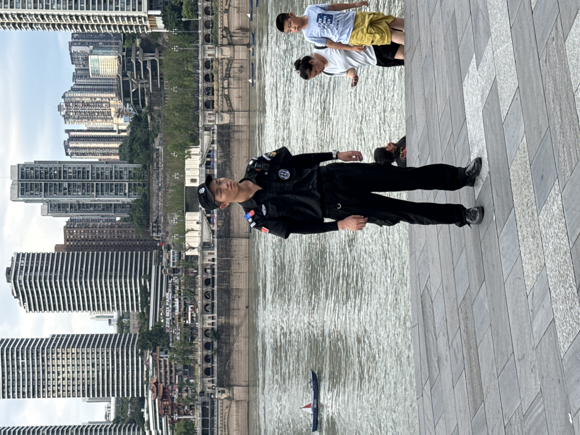 A man in a black uniform stands on a paved walkway next to a body of water, facing forward. He wears a black cap, black shirt with patches on the sleeves, and black pants. Behind him, across the water, is a city skyline with tall buildings and a waterfront promenade. To his right, a woman in a white shirt and black shorts and a boy in a blue t-shirt and yellow shorts stand looking towards the water. A small boat is visible on the water to the left of the frame. The sky is overcast. This scene is likely in Nan'an District, China, given the distinctive architecture.