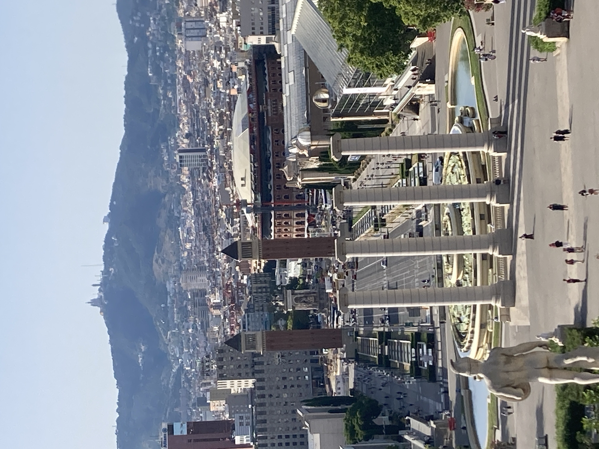 The image is a high-angle, vertical shot of a city skyline and a mountainous landscape. In the foreground, there is a large, ornate fountain with several statues and columns, and a pathway lined with people. Beyond the fountain, the city stretches out with numerous buildings of various sizes and architectural styles, densely packed. In the background, a large, tree-covered mountain rises, with more buildings scattered on its slopes and summit. The sky is clear and blue, suggesting a sunny day. The time of day appears to be late morning or early afternoon, indicated by the bright sunlight and shadows. The scene conveys a sense of urban vibrancy against a natural backdrop. Several people are visible walking along the paths and near the fountain, dressed in casual attire. A stone sculpture of a muscular figure is prominently positioned in the lower right foreground, facing away from the camera. The architecture of the buildings in the city suggests a European setting, and the context provided identifies the location as Barcelona, Spain. The overall impression is one of a well-developed city integrated with its natural surroundings. There is no visible text in the image.