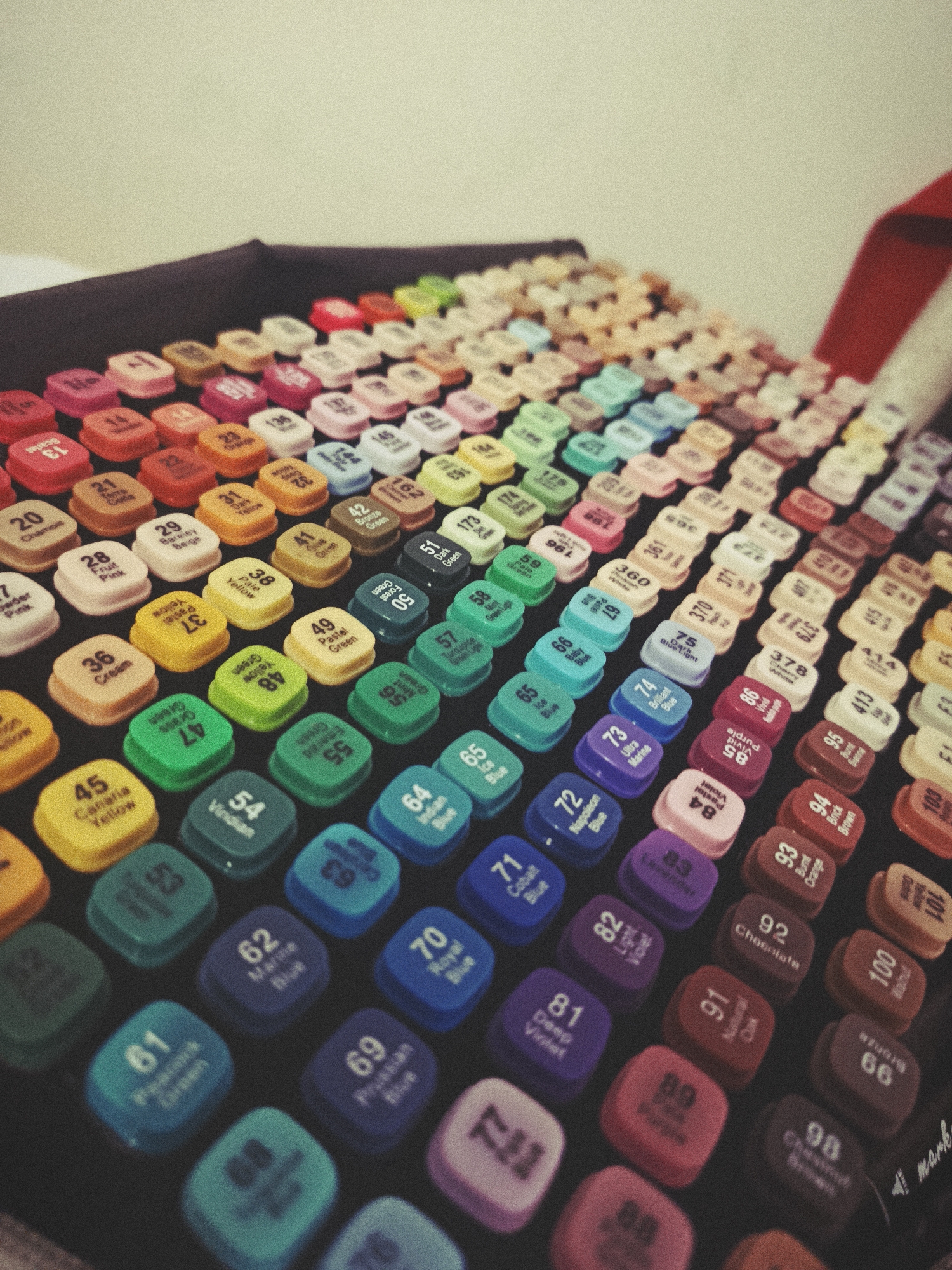 The image shows a collection of marker pen caps, arranged in rows on a dark surface. Each cap has a number and a color name printed on it. The markers are of various colors including yellow, orange, green, blue, pink, and purple. The image appears to be taken from a slightly elevated angle, with the markers filling the majority of the frame. The background is a plain, light-colored surface. There are no people or specific landmarks visible. The setting does not provide any indication of a specific location in the city of Hrodna, Belarus.
