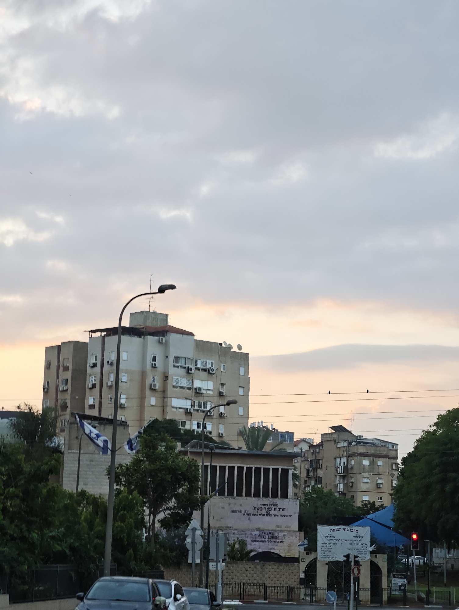 The image shows a street scene in Petah Tikva, Israel, at dusk. In the foreground, a street is visible with cars driving on it, and sidewalks with fences and vegetation. In the background, there are several buildings, including a tall apartment building and other multi-story structures. Two Israeli flags are visible, one on a flagpole near the apartment building and another attached to a pole further in the distance. There are also signs with Hebrew text visible on some of the buildings and near the street. The sky is cloudy, with soft light suggesting either sunrise or sunset. The overall impression is a typical urban environment.