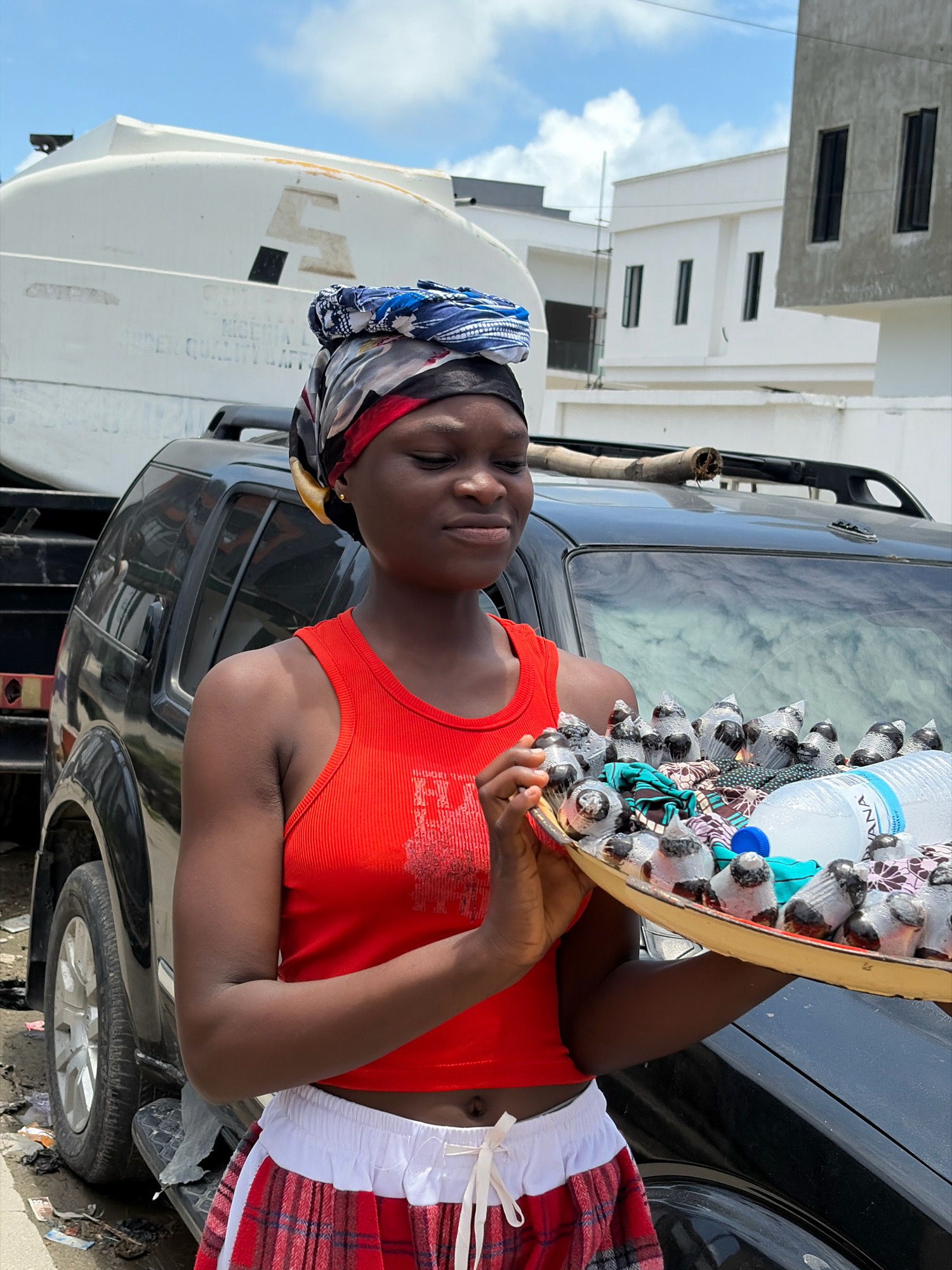 A young Black woman with dark skin, wearing a red tank top, a colorful headwrap, and a plaid skirt, stands in what appears to be a residential area in Igboefon, Nigeria. She is holding a tray filled with small, individually wrapped items, possibly snacks or candies, and a large bottle of water. The background shows a large white vehicle with some text on its side, and modern white buildings. She is positioned in front of a black SUV. The sky is partly cloudy.