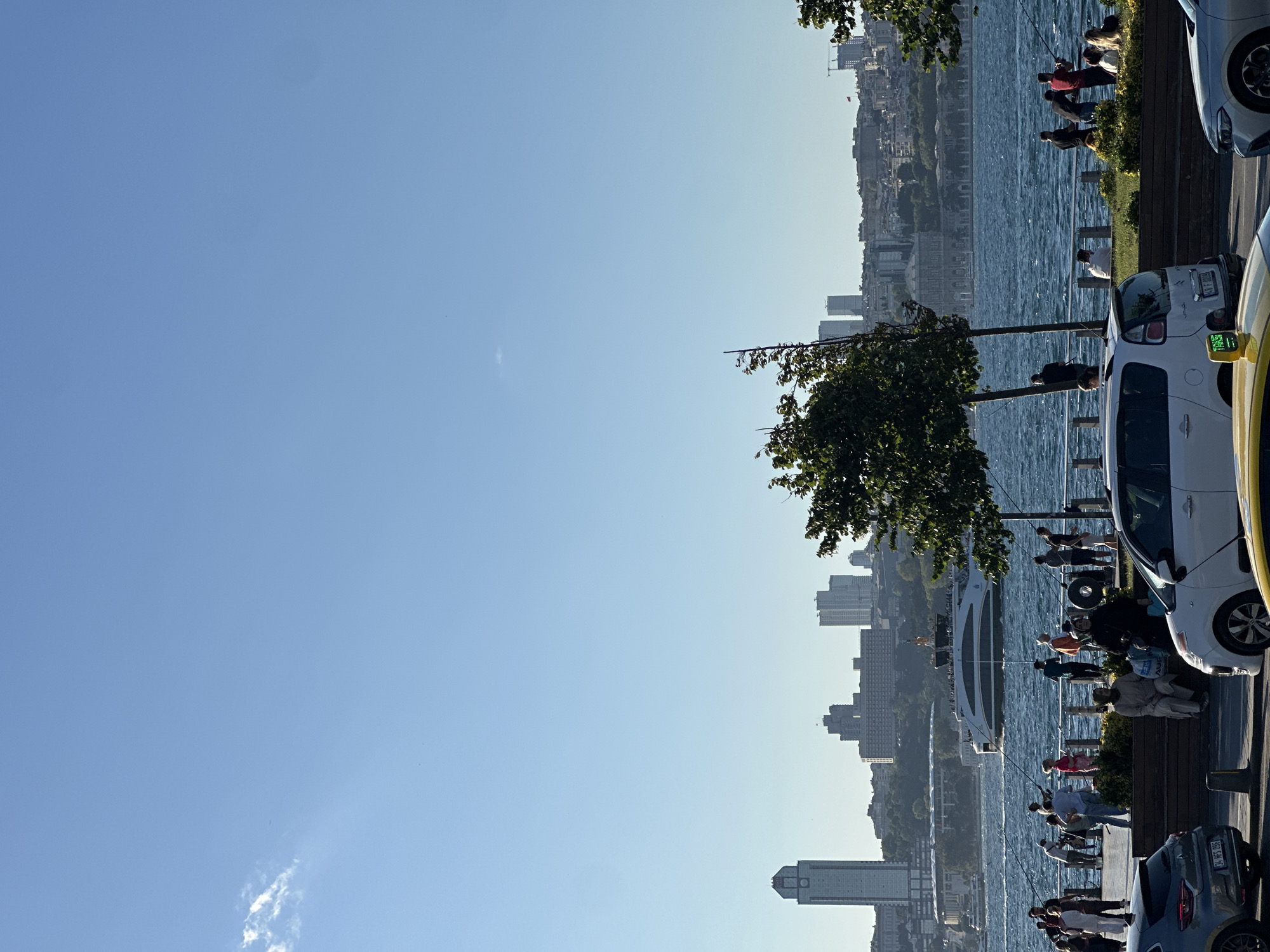 The image depicts a sunny day in Üsküdar, Turkey, with a clear blue sky dominating the upper portion. In the foreground, a street runs parallel to a waterfront promenade. Several cars are parked along the street, including a grey Renault with a Turkish license plate and a white compact car, which appears to be a taxi with a "TAXI" sign on its roof. A portion of a silver car is also visible on the right.
The promenade is populated with numerous people. Some are walking, while others are seated on benches or standing by the railing. A group of individuals are actively engaged in fishing with long rods, casting their lines into the shimmering blue water. Another person appears to be walking a dog.
Across the water, a large ferryboat is underway, carrying passengers. The background showcases the cityscape of Istanbul, characterized by a collection of buildings, including several modern skyscrapers and older structures. The water itself is moderately choppy, suggesting a gentle breeze. Trees with green foliage are interspersed along the promenade, providing some shade. The overall scene conveys a leisurely atmosphere on a bright day by the Bosphorus strait.