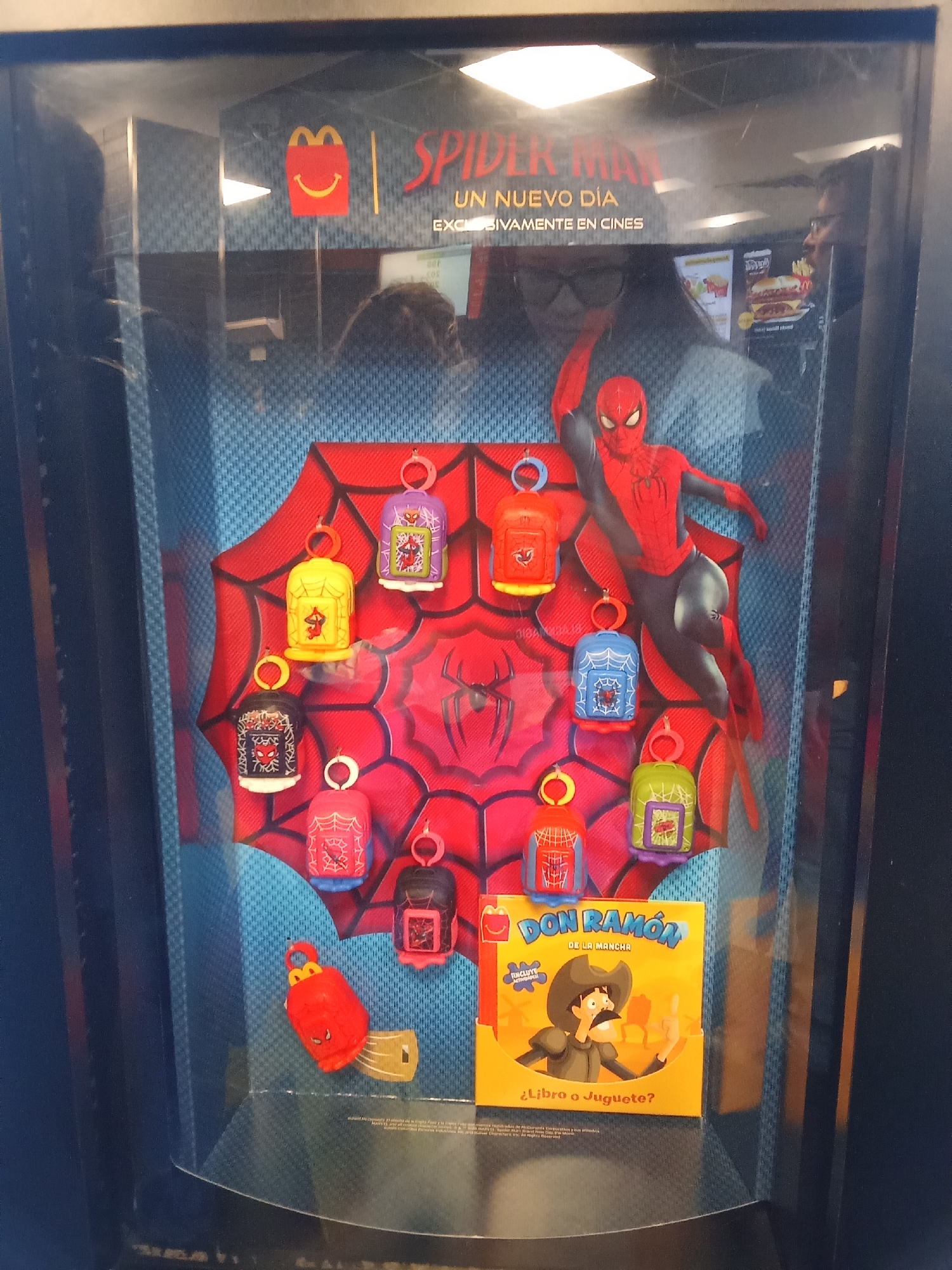 The image shows a display case at a McDonald's restaurant in Santiago, Chile, featuring toys from the movie "Spider-Man: Un Nuevo Día". The display case is made of glass and has a blue, patterned background with a large image of Spider-Man in the center. Various small, bell-shaped toys with keychains, each depicting a different design related to Spider-Man or the McDonald's Happy Meal, are arranged around the central image. One toy is a bright red spider-shaped figure. At the bottom of the display is a yellow box featuring the character "Don Ramón de la Mancha" and the text "¿Libro o Juguete?", implying it's a promotional item. Reflections in the glass show the interior of the McDonald's, including other customers and menu boards.