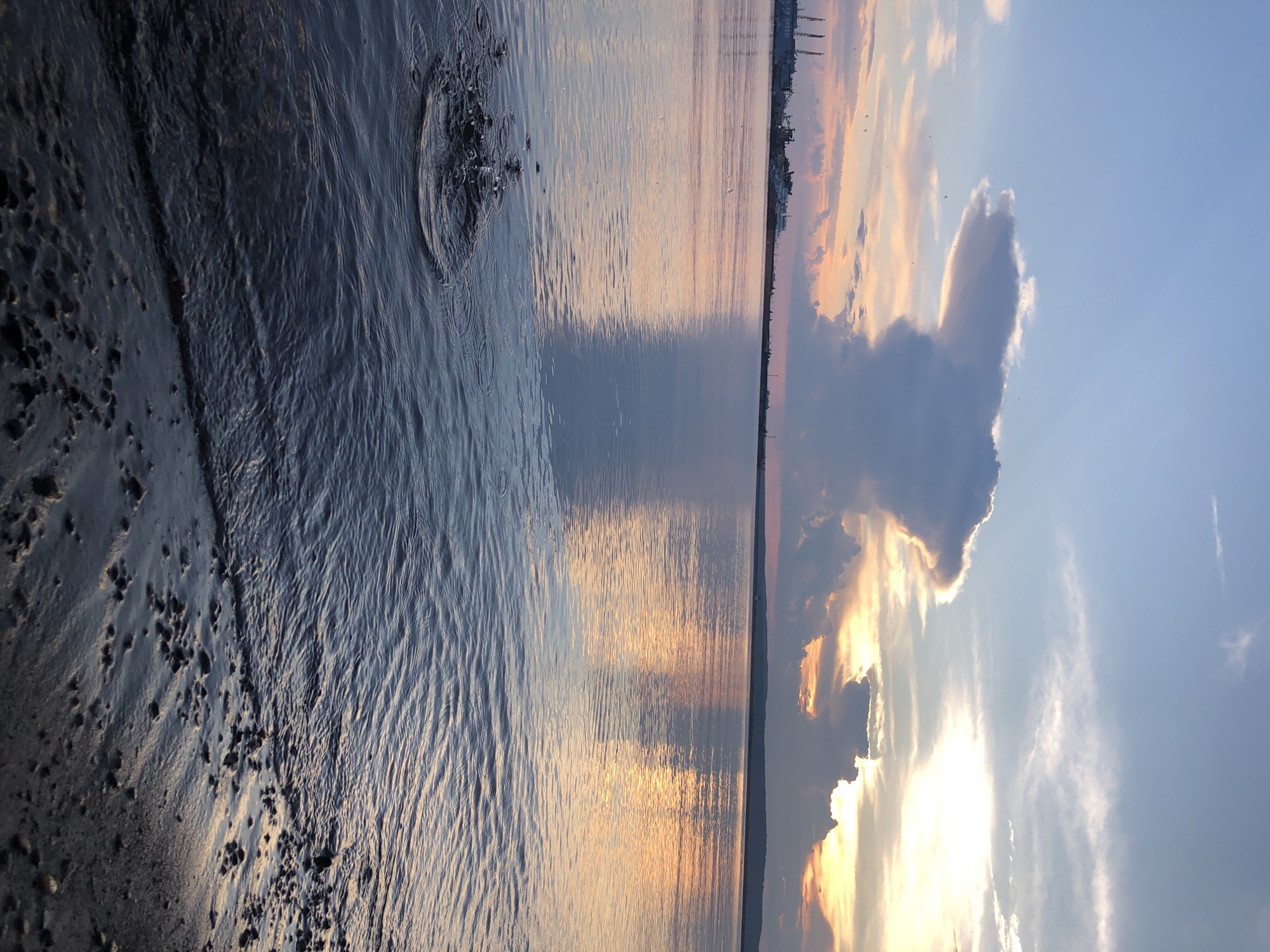 The image depicts a sunset over the Kama River in Chaikovsky, Russia. The sky is a gradient of blue, pink, and orange, with dramatic clouds casting shadows on the water. In the foreground, ripples on the water's surface indicate a recent splash. The shoreline is visible on the right, composed of wet sand and small pebbles. In the distance, industrial cranes and structures dot the horizon. The overall atmosphere is tranquil and serene, capturing the natural beauty of the river at dusk.
