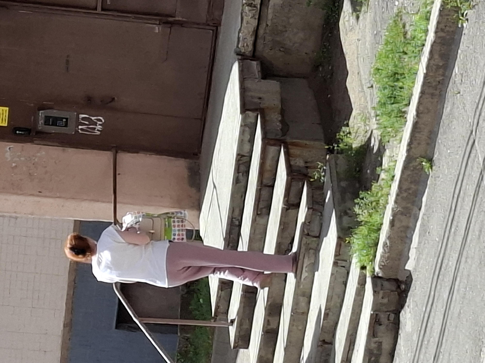 A person with red hair tied up is walking up a set of concrete stairs. They are wearing a white t-shirt and pink pants. The person is carrying a bag over their left shoulder and a light-colored tote bag in their right hand. The stairs are worn and have some grass growing around them. Behind the stairs is a brown metal door with a gray intercom panel and a label with a symbol. The area appears to be an outdoor urban setting.