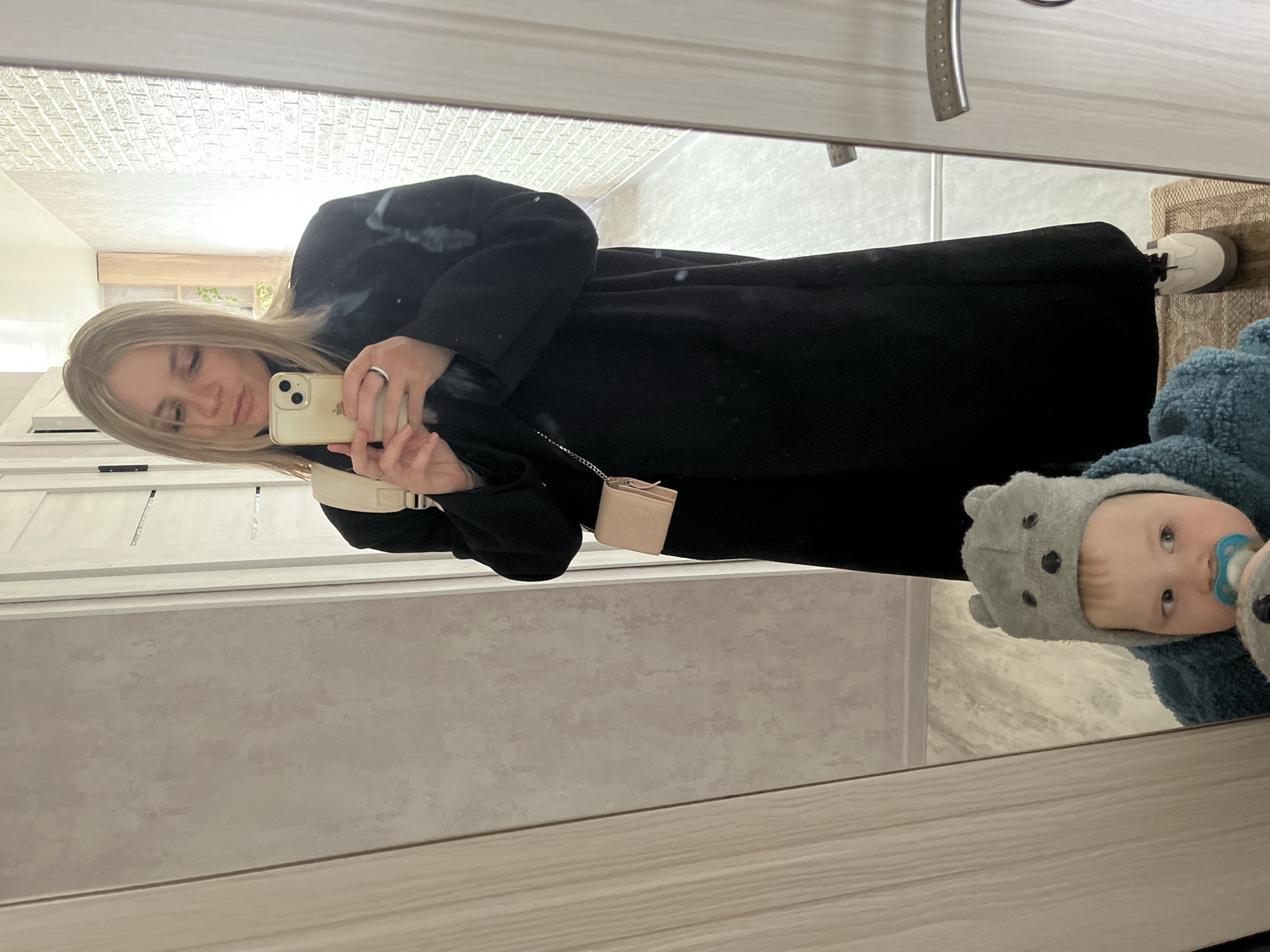 The image, rotated 90 degrees clockwise in its original display, captures a mirror selfie of a woman and a child in an indoor setting in Chaikovsky, Russia.
The main subject is a woman with long blonde hair, wearing a black long coat or dress. She holds a gold-colored smartphone, likely an iPhone due to its distinct camera bump, in her right hand to take the selfie. A silver ring is visible on her left ring finger, and a small, light-colored cross-body bag with a chain strap is worn over her shoulder. Her expression is neutral to slightly smiling.
Reflected in the lower right portion of the mirror is a baby or toddler. The child wears a blue, fuzzy garment and a grey hat with bear ears. A blue pacifier is in their mouth, and they appear to be looking directly into the mirror.
The background, reflected in the mirror, shows a brightly lit room. Visible details include a textured white ceiling (possibly patterned wallpaper or ceiling tiles), light-colored walls, and a light wooden door frame or trim. In the reflection's upper left, a light-colored wooden shelf or cabinet with some green plant foliage is visible. A silver door handle is partially seen on the right side, and a beige, textured mat or rug is discernible at the bottom right. The overall impression is a clean, simple, and well-lit indoor space, likely a hallway or room with a large mirror. There is no visible text in the image.