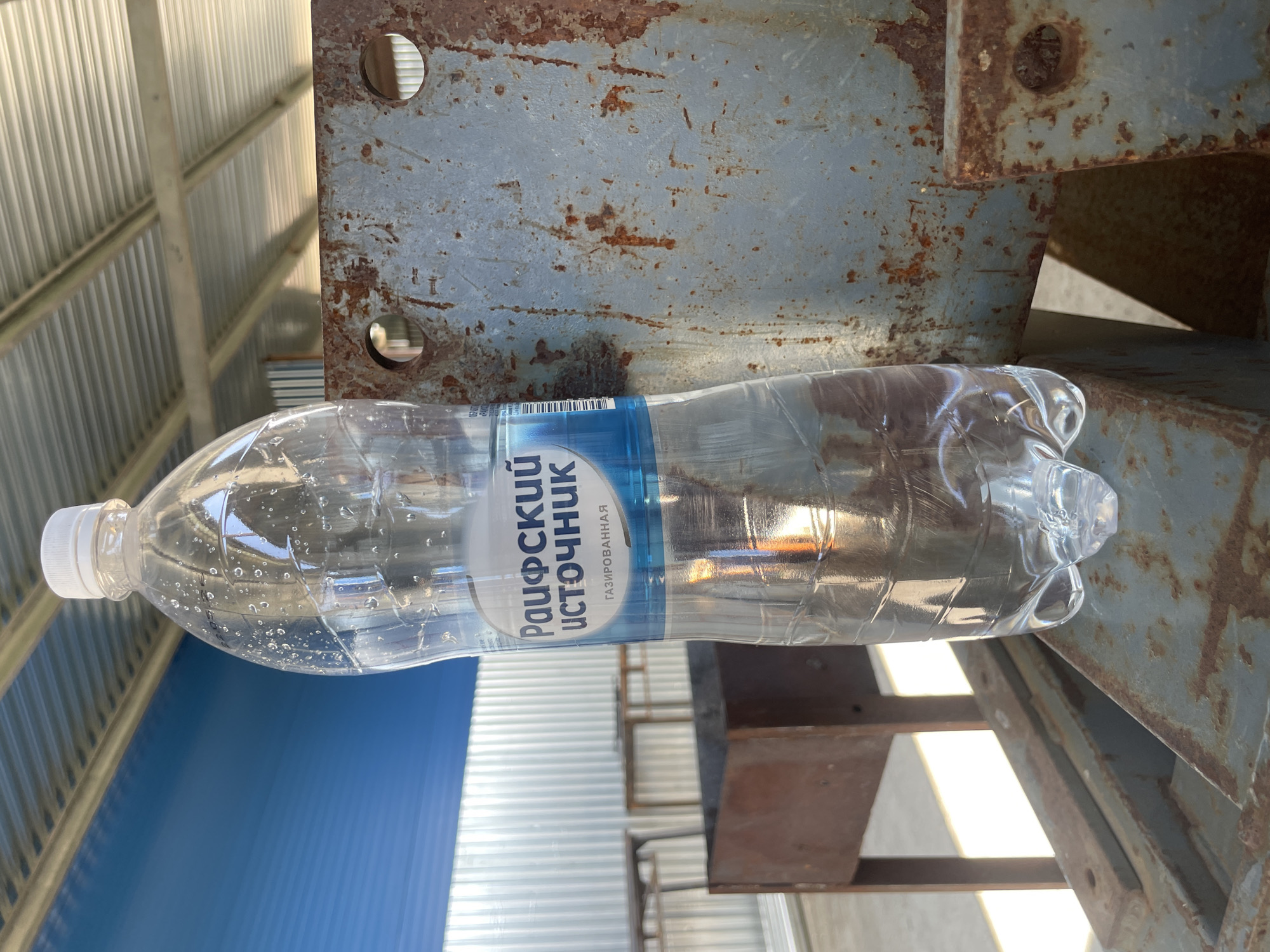 A clear plastic bottle of "Raufoa 's Source" brand carbonated water is positioned on a rusty metal surface. The bottle is half-full, with condensation visible on its exterior. In the background, a corrugated metal roof is supported by beams, and a blue wall is visible to the left. Further back, metal tables and a barbecue grill are present, suggesting an outdoor recreational area, likely a dacha or picnic spot, potentially in the village of Sokury, Russia. The lighting indicates daylight, with sunlight reflecting off the water bottle and creating shadows.