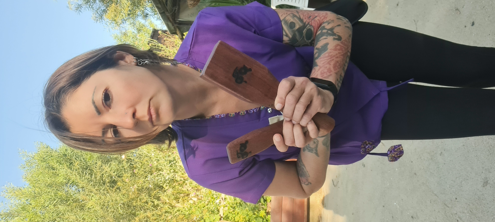 A person is shown in a close-up, outdoor portrait, looking directly at the camera. She has medium-brown hair, dark eyes, and several facial piercings including a nose stud, a septum ring, and decorative earrings. She is wearing a purple short-sleeved top with a vibrant, multi-colored patterned trim along the neckline and a matching tie detail visible on her side. Black pants complete her attire.
Both forearms are heavily tattooed with dark, intricate designs; the left arm features mechanical-themed imagery, possibly pistons, while the right arm displays complex patterns. A black smartwatch is worn on her left wrist. She holds two flat, rectangular wooden paddles, one in each hand. Each paddle is dark-stained wood and features a distinctive, etched or burned black symbol, resembling a stylized animal such as a pig or boar.
The background consists of a clear blue sky and abundant green foliage from trees or bushes, suggesting a natural, open-air setting. The ground is a light-colored paved surface, possibly concrete. The scene takes place during daytime, under bright, natural light in the Lachish Regional Council, Israel. The person's expression is neutral and engaging. No visible text is present in the image.