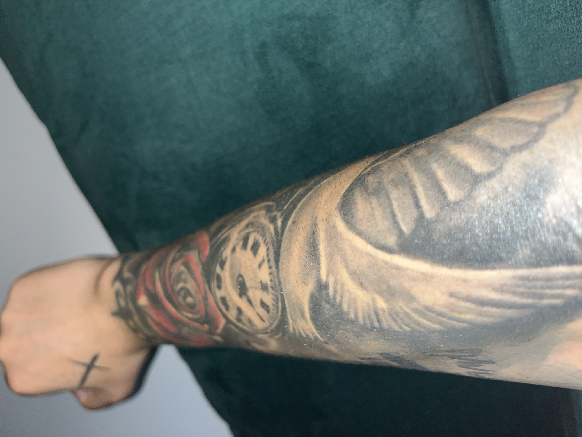 An arm with tattoos is resting against a dark green surface. On the wrist, there is a tattoo of a rose with red and black shading. Above the rose, closer to the hand, there is a tattoo of a clock face with Roman numerals. Further down the arm, there are blurred images of what appear to be feathers or wings. A small black cross tattoo is visible on the back of the hand. The background is out of focus and consists of a light-colored wall and the dark green surface.
