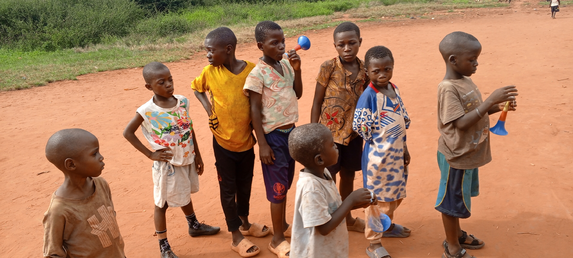 This outdoor photograph captures nine young boys gathered on a reddish-brown dirt path in Ngalula, Democratic Republic of the Congo. The setting features lush green vegetation in the background, indicative of a rural or semi-rural environment. The bright, clear daylight suggests it is mid-day or afternoon.
The boys, ranging from childhood to early adolescence, have closely cropped dark hair and are dressed in various casual, worn clothing, including t-shirts, sleeveless tops, and shorts. Some wear sandals or flip-flops, while one has dark shoes. On the far left, a boy in a light brown t-shirt looks right. Next to him, a boy in a white sleeveless top with colorful patterns and the visible text "TRAVEL TIME" stands with hands on his hips. Another boy in a yellow short-sleeved shirt also has his hands on his hips.
Several children are engaged with plastic toy horns. One boy in a light patterned shirt holds a blue and red horn to his mouth, while another on the far right, wearing a brown t-shirt and blue shorts, holds a multi-colored (blue, orange, yellow) horn in both hands. In the center, a boy in a blue and white patterned shirt holds a small object in his hand. Their expressions are generally neutral or contemplative, suggesting an informal gathering or a pause during play. A partial view of "EURO20" is visible on one boy's shorts.