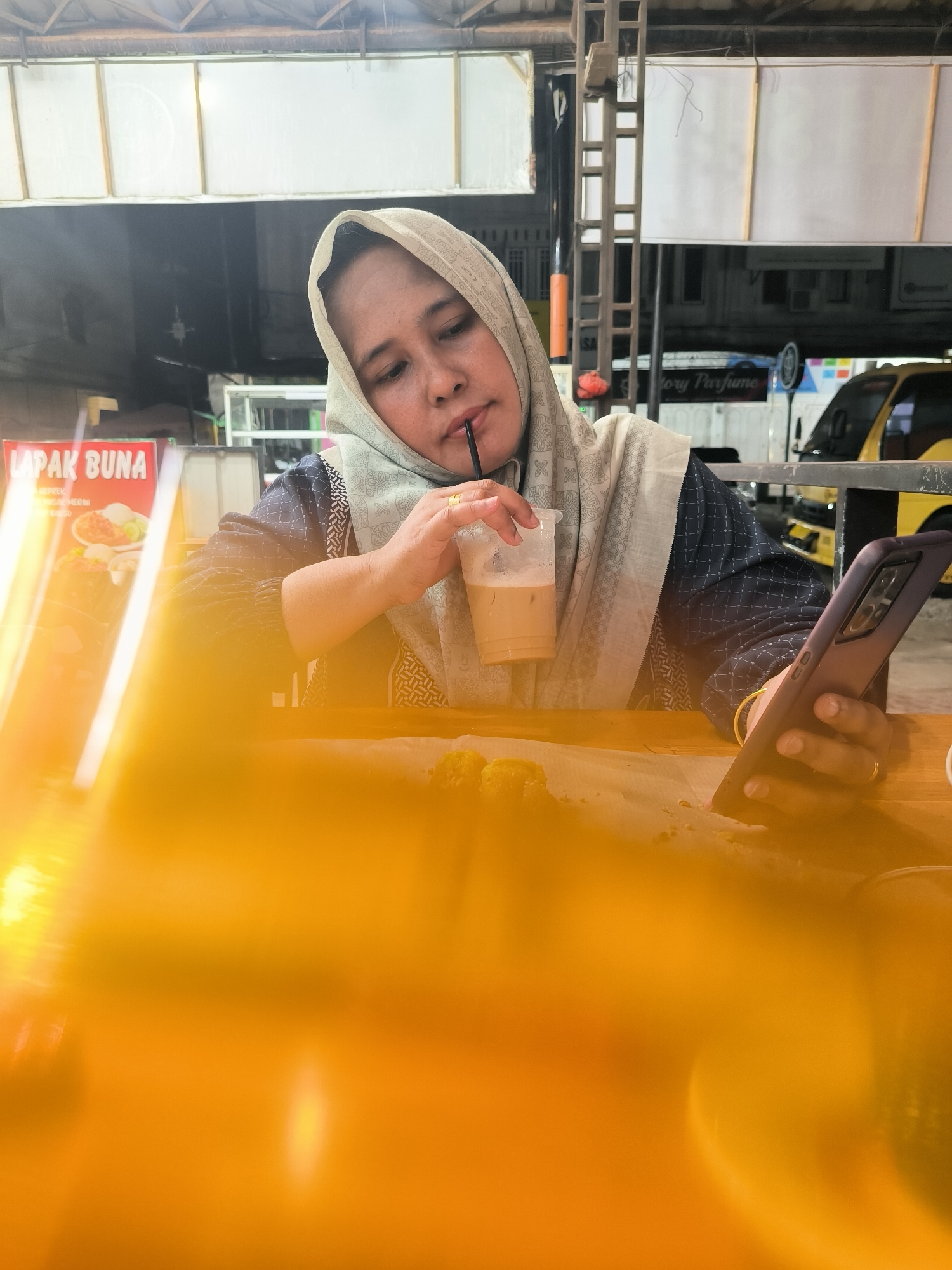 The image features a woman in a hijab sitting at an outdoor table and looking at her phone while drinking a beverage. The setting appears to be a street-side establishment.
The woman is wearing a hijab. She is holding a phone in her right hand. In her left hand, she holds a clear plastic cup with a straw, containing a light-brown liquid.
On the table, some small, round food items are visible. Behind the woman, in the background, are a building facade, a sign reading "LAPAK BUNA," and a vehicle. Another sign "tory Perfume" can also be seen.
The image was taken in Lhokseumawe, Indonesia.