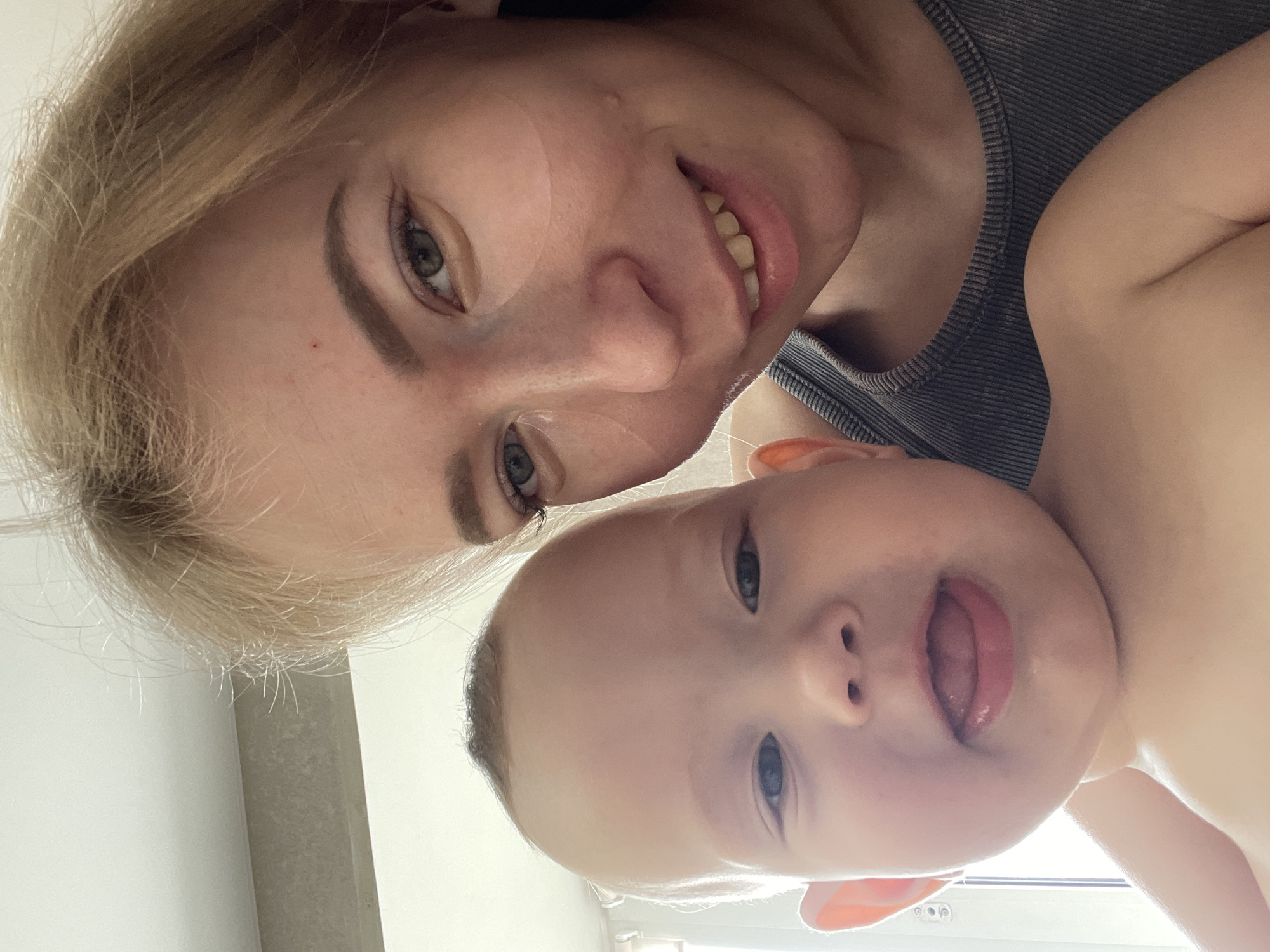 A close-up selfie shows a woman and a baby. The woman has blonde hair, fair skin, and green eyes. She is smiling. The baby is fair-skinned with blue eyes and has their tongue partially sticking out. The woman is wearing a gray tank top. The background is blurred and indistinct. There is no specific information to locate the image in the city of Perm, Russia.