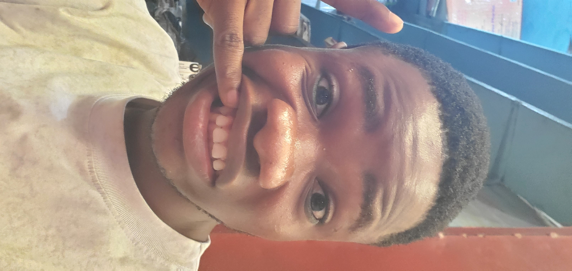 A close-up, rotated portrait shows a person with dark skin and short, curly black hair. The individual is making a playful or exaggerated facial expression, using their right index finger to pull up their upper lip, revealing their upper teeth and gums. Both eyes are open and looking towards the camera. The person is wearing a light-colored (possibly white or very light yellow) t-shirt, which has a ribbed collar and a faint white mark on the fabric. The skin appears somewhat shiny, possibly from sweat or oil. The background is out of focus, featuring indistinct shapes including horizontal lines that could be shelves or structural elements, and a reddish surface towards the bottom right. Faint, partially visible letters "e" and "o" are discernable on a white background in the blurry upper-mid portion of the image, potentially part of a sign or label.