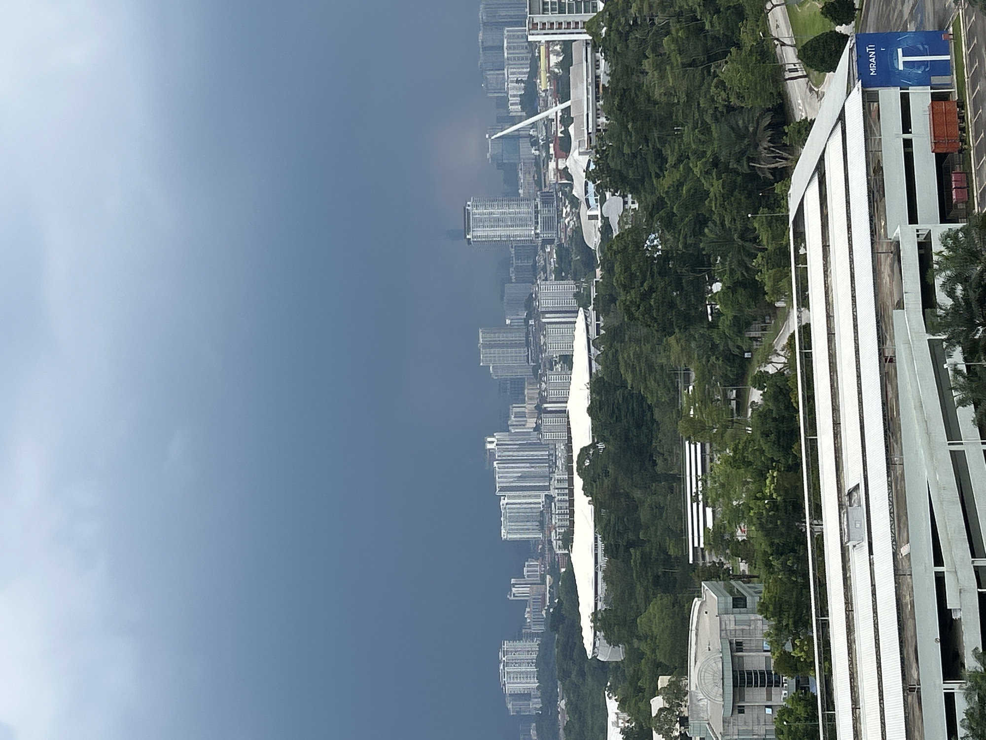The image presents an elevated view of Kuala Lumpur, Malaysia, under a cloudy sky. The cityscape features numerous high-rise buildings of varying architectural styles, densely packed in the central area. Lush green trees are interspersed among the buildings, particularly along the fringes of the developed area. In the foreground, a large white building with vertical lines dominates the right side of the frame, with a blue banner displaying the word "MIRANTI". A road and parking area are visible next to this building. The environment is urban, with a blend of natural and built elements.