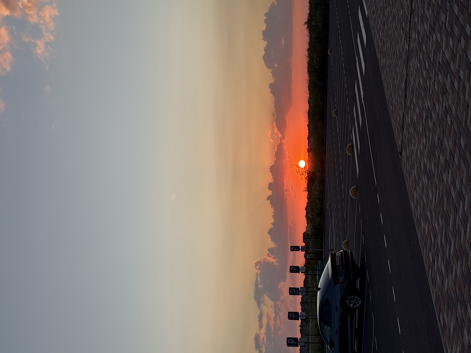 A car is parked in an empty parking lot at sunset. The sky is a gradient of orange and pink, with scattered clouds. The sun is a bright, round orb low on the horizon. The parking lot is paved with asphalt and has white lines marking the parking spaces. There are yellow bollards between some of the spaces. On the right side of the image, there is a brick walkway. In the background, there are trees and a few buildings. The scene appears to be in Mikhaylovsk, Russia. The overall mood is peaceful and serene. There is no visible text in the image.