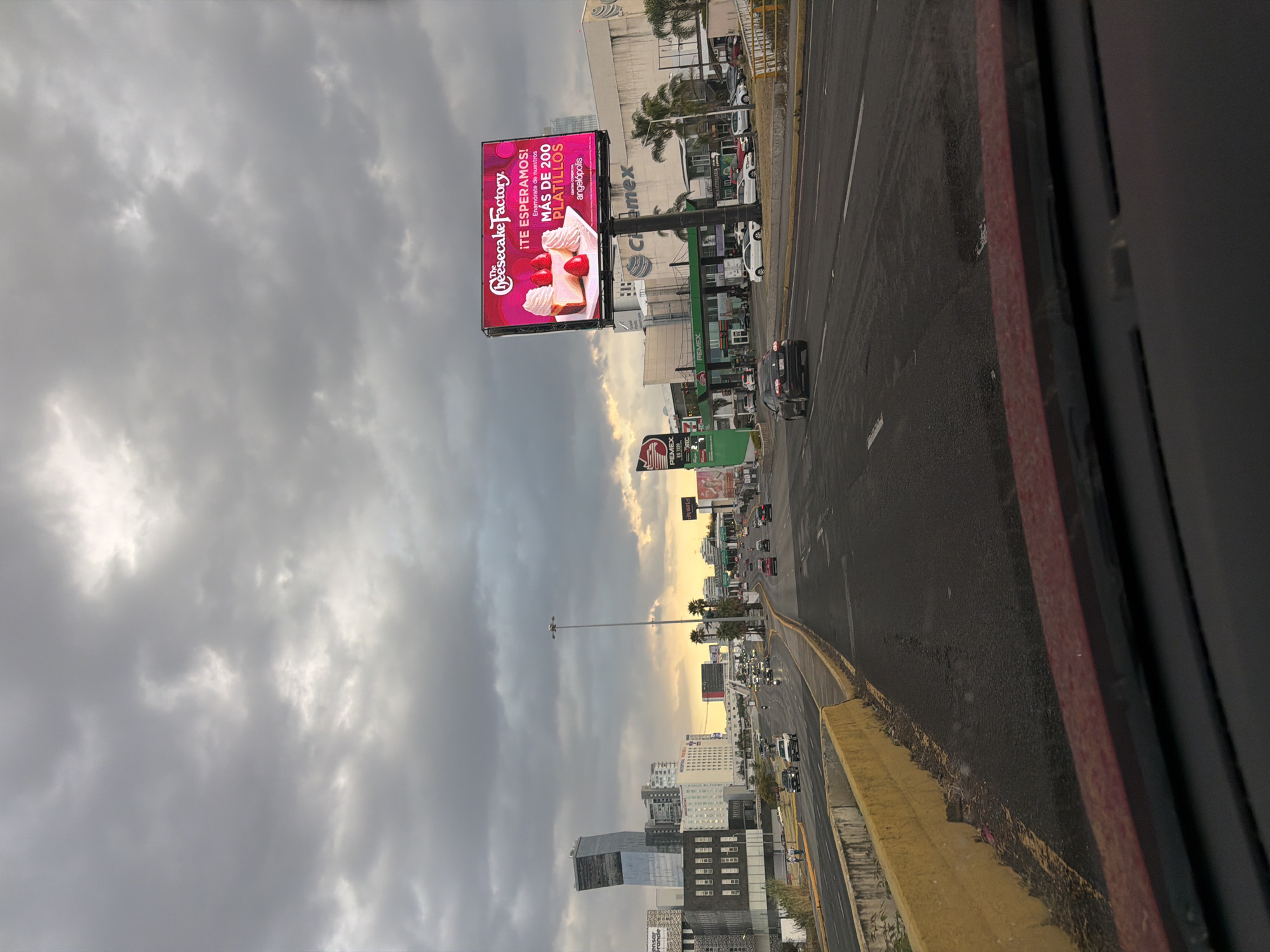 The image shows a street scene in Puebla, Mexico, with a focus on a large billboard for The Cheesecake Factory. The billboard advertises over 200 menu items and refers to "angelópolis," which is a well-known area of the city. Below the billboard is a Pemex gas station. The street has traffic, including cars and a truck. The sky is cloudy, with a hint of sunset. Tall buildings and palm trees are visible in the background. The photo was taken from an elevated position, likely inside a vehicle, as part of the window frame is visible.
