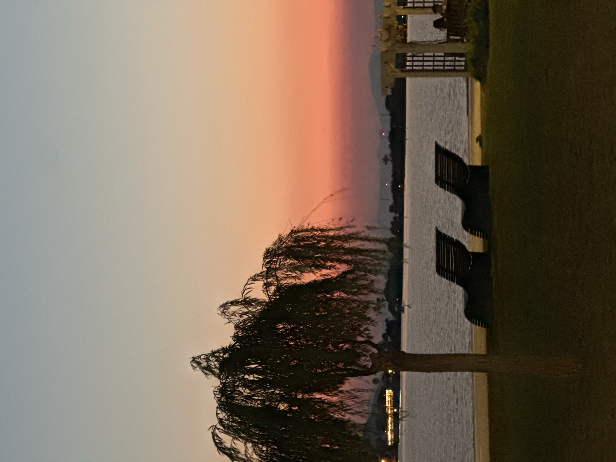 The image depicts a serene sunset scene at a waterfront location, likely in Fethiye, Turkey. The sky displays a gradient of colors, transitioning from a pale blue at the top to a warm orange and pink near the horizon. In the foreground, a weeping willow tree stands on a grassy area, its branches drooping. Two dark-colored lounge chairs are positioned on the sand near the water's edge. To the right, a white gazebo structure is visible with a few people seated within it. The water is calm, reflecting the soft light of the sunset. In the background, distant hills are silhouetted against the colorful sky, and lights from buildings are faintly visible across the water. The overall atmosphere is peaceful and tranquil, suggesting an evening setting.