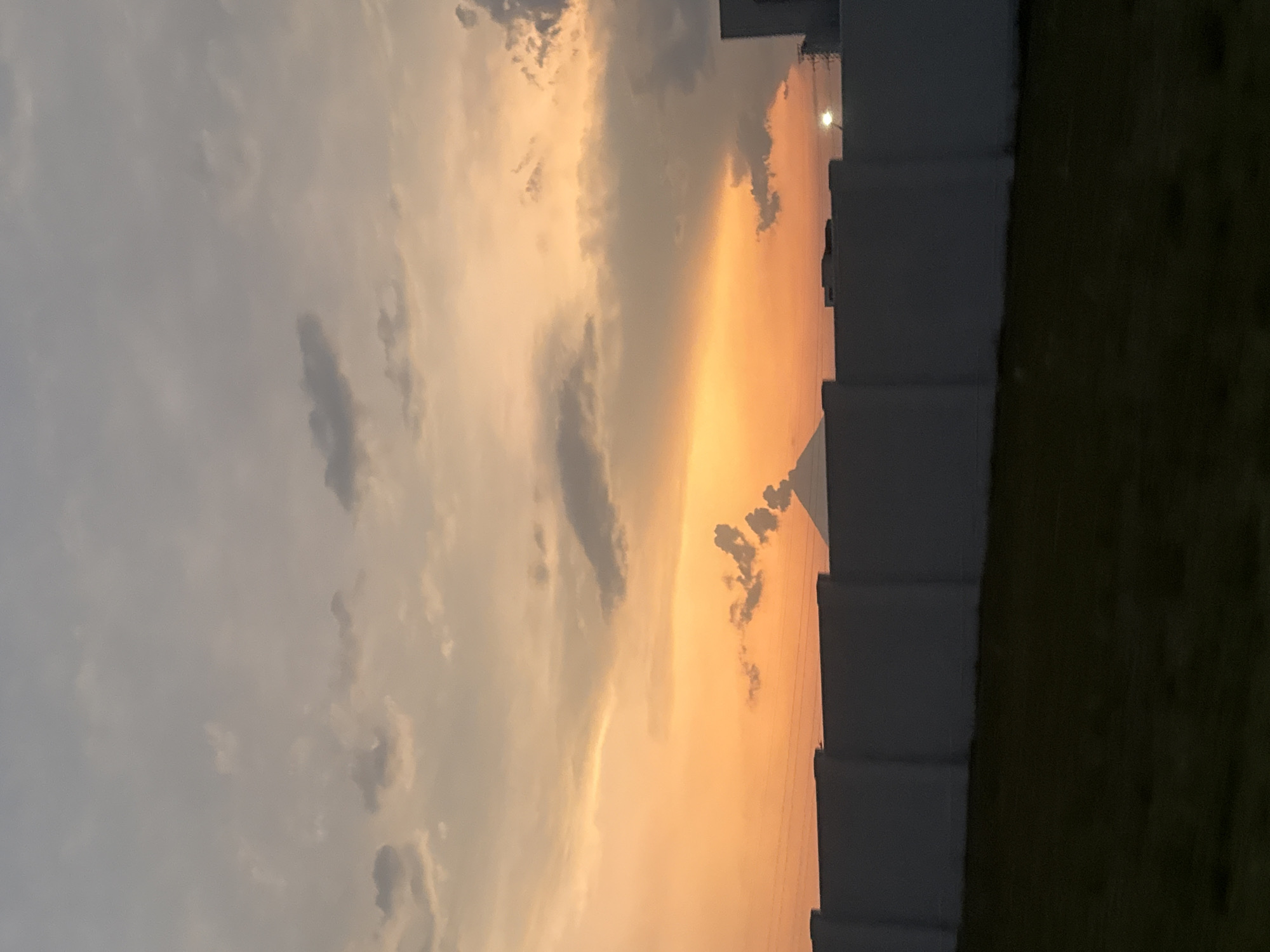 The image shows a view of the sky during sunset, with colors ranging from gray to orange. The left side of the frame is dominated by a cloudy sky. On the right, a dark-colored building with a blocky facade is visible, along with some dark green grass. The location is speculated to be the city of San Bernardino Tlaxcalancingo, Mexico.