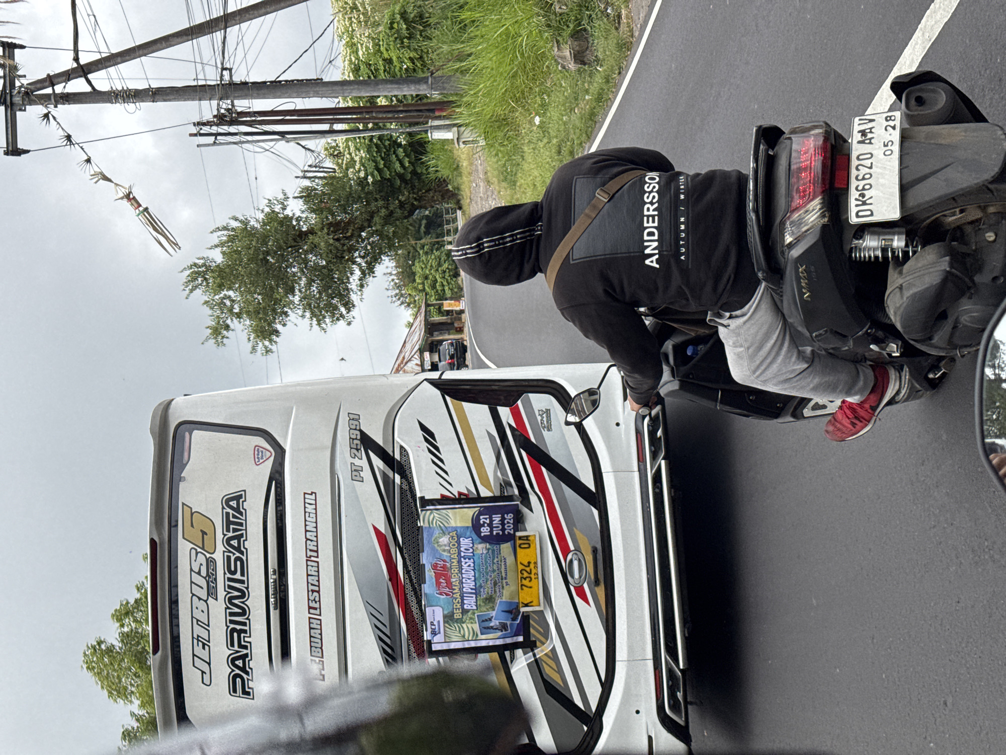 The image shows a street scene in Kintamani, Indonesia, on a cloudy day. A white tour bus with "JETBUS 5 PARIWISATA" written on its side is in the foreground, displaying a promotional poster for a "BALI PARADISE TOUR" scheduled for June 18-21, 2026. To the right of the bus, a person wearing a black hoodie with "ANDERSSON" and "AUTUMN / WINTER" printed on it, rides a black scooter. The scooter has a license plate reading "DK 6620 AAV 05-28". The road is paved and has a white line marking the lane. Lush green vegetation and trees are visible on the right side of the road, with power lines and poles overhead. In the distance, a small building with a thatched roof can be seen. The overall atmosphere is one of everyday life in a roadside setting.
