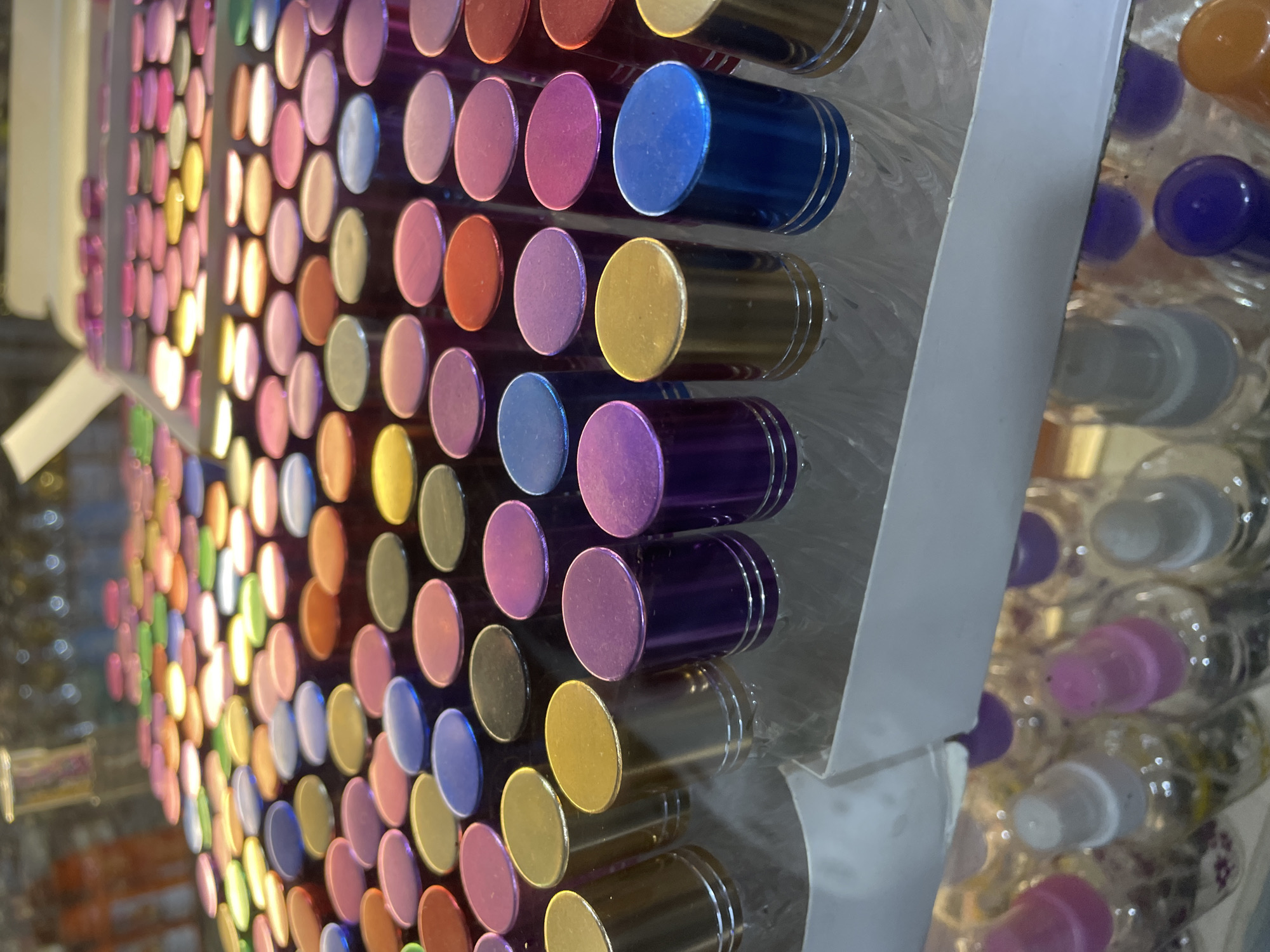 The media file shows a close-up, angled view of a display of colorful lipstick tubes arranged in rows. The lipsticks have metallic caps in various shades of pink, purple, blue, gold, and red, with some caps appearing iridescent. They are presented vertically in a grid-like fashion, suggesting a retail or promotional setting. The background is blurred but appears to be a retail environment with other displays and lighting. The focus is on the vibrant colors and metallic finishes of the lipstick caps. There is no visible text in the image. The setting is likely a store in North Jakarta, Indonesia, given the context. The image captures a moment of retail display, highlighting the aesthetic appeal of the cosmetic products.