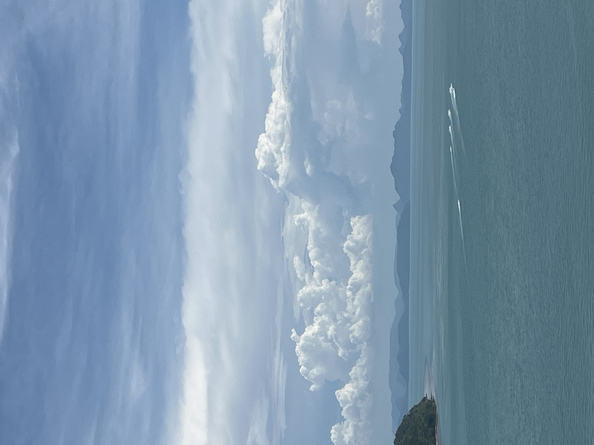 This is a wide-angle, eye-level shot of a turquoise ocean scene under a partly cloudy sky, likely captured in Baan Thong Krut, Thailand. The foreground is dominated by the tranquil, rippling surface of the sea. In the mid-ground, a cluster of speedboats are visible, leaving trails of white wake as they move from left to right across the frame. To the far left, a lush green, tree-covered island juts into the water. In the distance, a hazy range of blue mountains stretches across the horizon, separating the sea from the sky. The sky itself is a mix of light blue and white clouds, with some denser, cumulonimbus formations adding texture. The overall impression is one of a bright, clear day in a tropical coastal setting.
