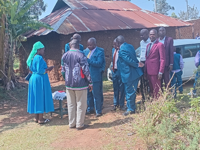A group of people stand outside a small mud house with a rusty corrugated metal roof. Most men wear suits in shades of blue or maroon. A woman in a bright blue dress and headscarf stands to the side, near a small table covered with a white lace cloth. A white car is parked nearby. Some individuals are engaged in conversation. The setting appears to be a rural area in Africa.