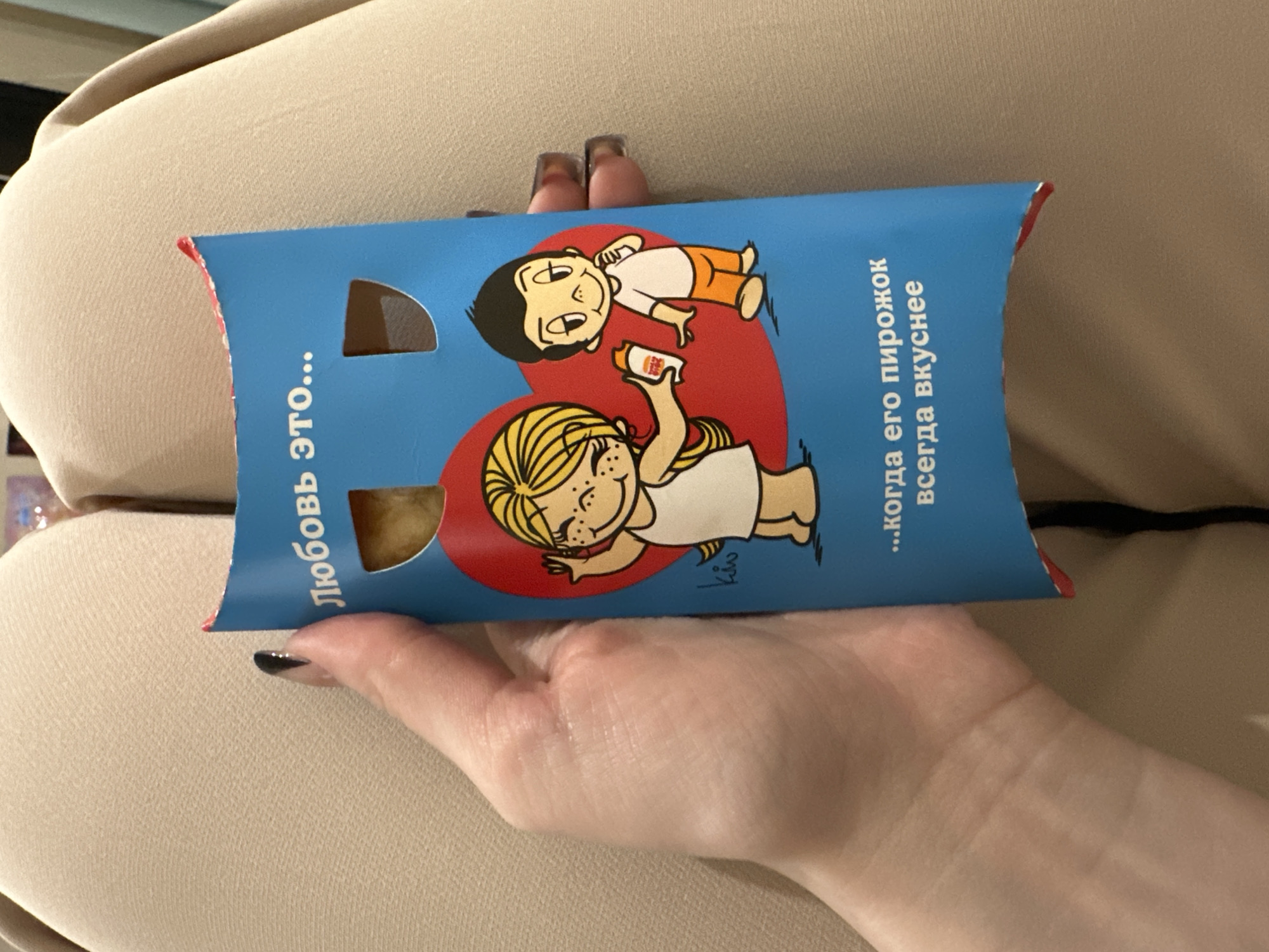 A hand holds a blue cardboard box with a red heart design featuring cartoon figures. The box has two die-cut openings revealing the contents, which appear to be pastries. Text in Cyrillic is printed on the box. The box is positioned against a background of beige fabric. The location is not specified.