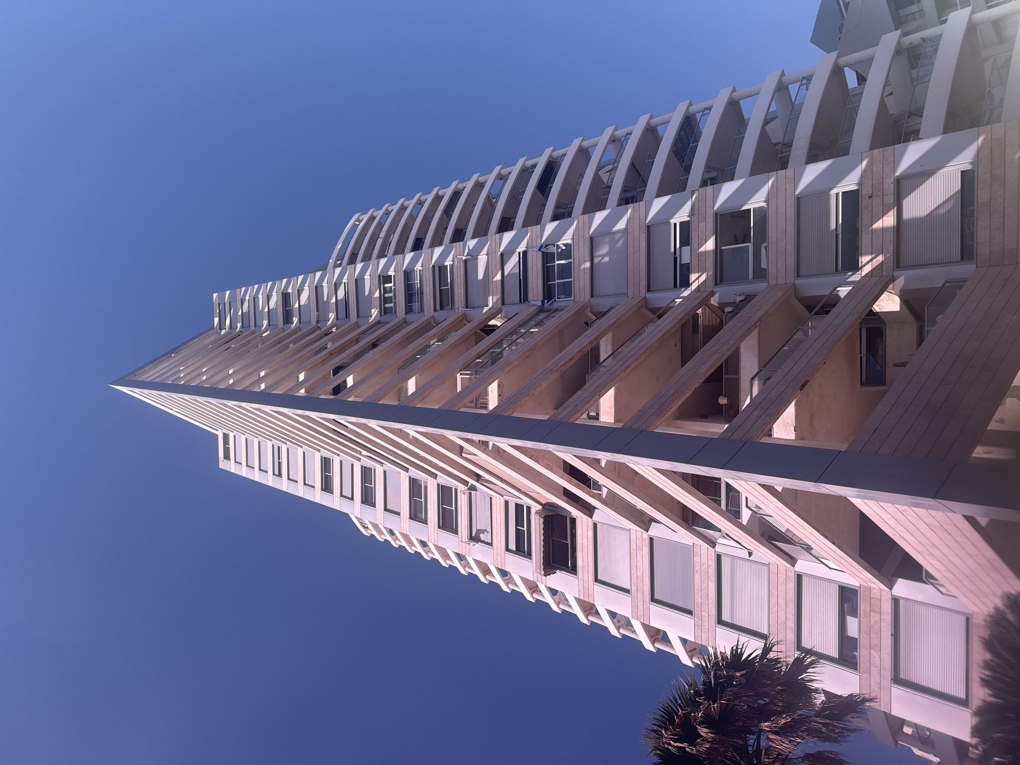 A low-angle, upward-looking shot captures the imposing modern architecture of a building against a clear, vibrant blue sky. The perspective creates a dynamic view, making the tall structure appear to lean diagonally across the frame.
The building is characterized by its distinctive design. The upper section features a series of prominent, curved, white arch-like structures that protrude from the facade, resembling ribs or an artistic skeletal framework. These arches cast strong, angular shadows onto the lighter-toned building surface below. Beneath these arches, multiple rows of windows are visible, many equipped with closed roller blinds or shutters, suggesting residential or office units.
Further down the building, a different architectural style emerges, with visible horizontal beams and diagonal supports creating a layered and textured facade. The materials appear to be light-colored, possibly stone or concrete, which glow softly in the bright sunlight.
The clear blue sky indicates fair weather, and the sharp, well-defined shadows suggest strong sunlight, likely from midday or early afternoon. In the bottom right corner of the image, the fronds of a palm tree are partially visible, adding a natural element to the urban landscape and hinting at a warm climate. No human activity or visible text is present in the image. The scene is set in Bat Yam, Israel.