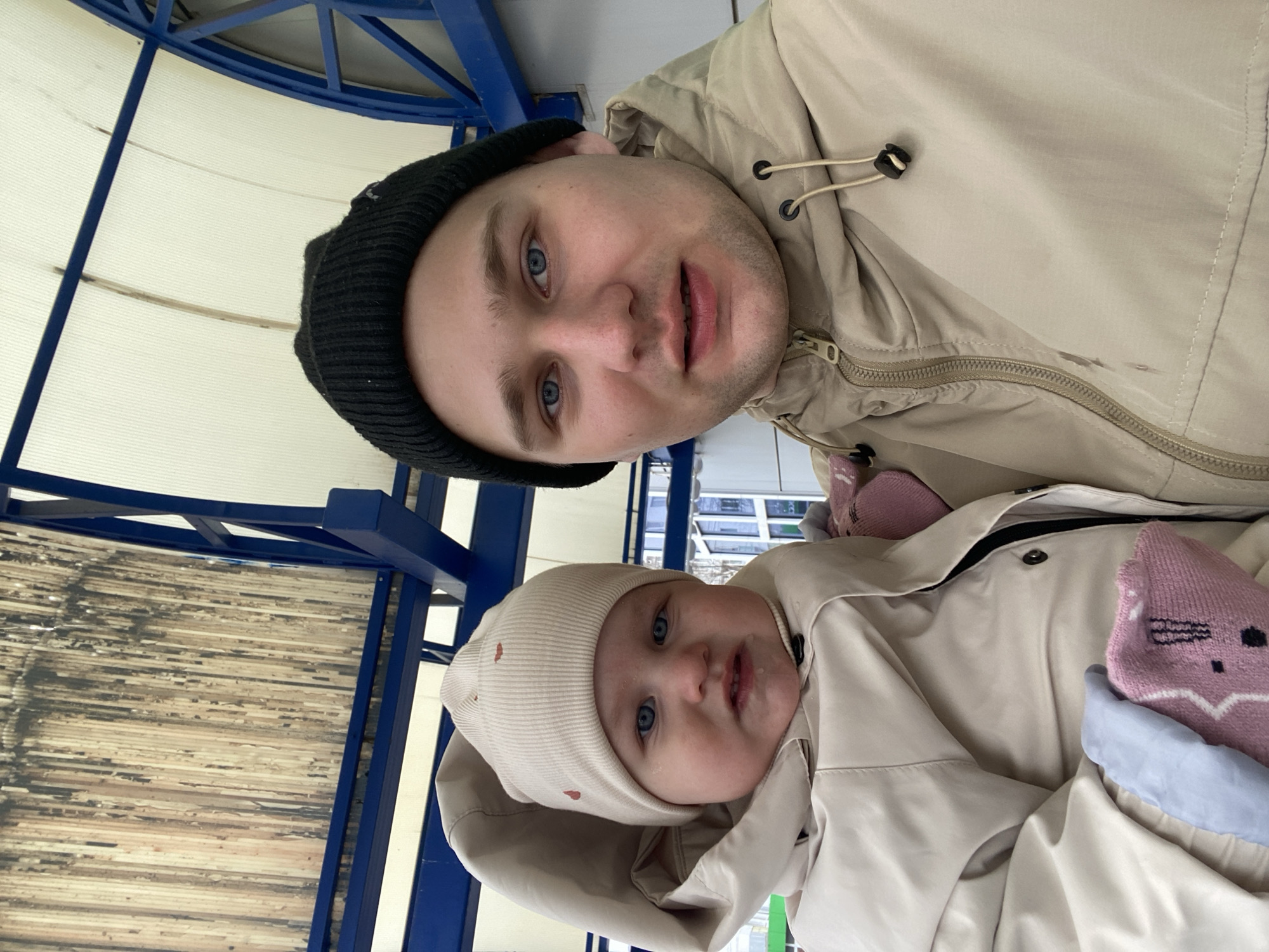 A selfie photograph shows a man and a baby outside on a partly sunny day. The man wears a black knit cap and a tan hooded jacket. The baby wears a tan hooded jacket and a matching knit hat. A pink and white animal-themed sock is visible near the baby. A blue-painted metal structure supports a weathered wood ceiling. The location is Chaikovsky, Russia.